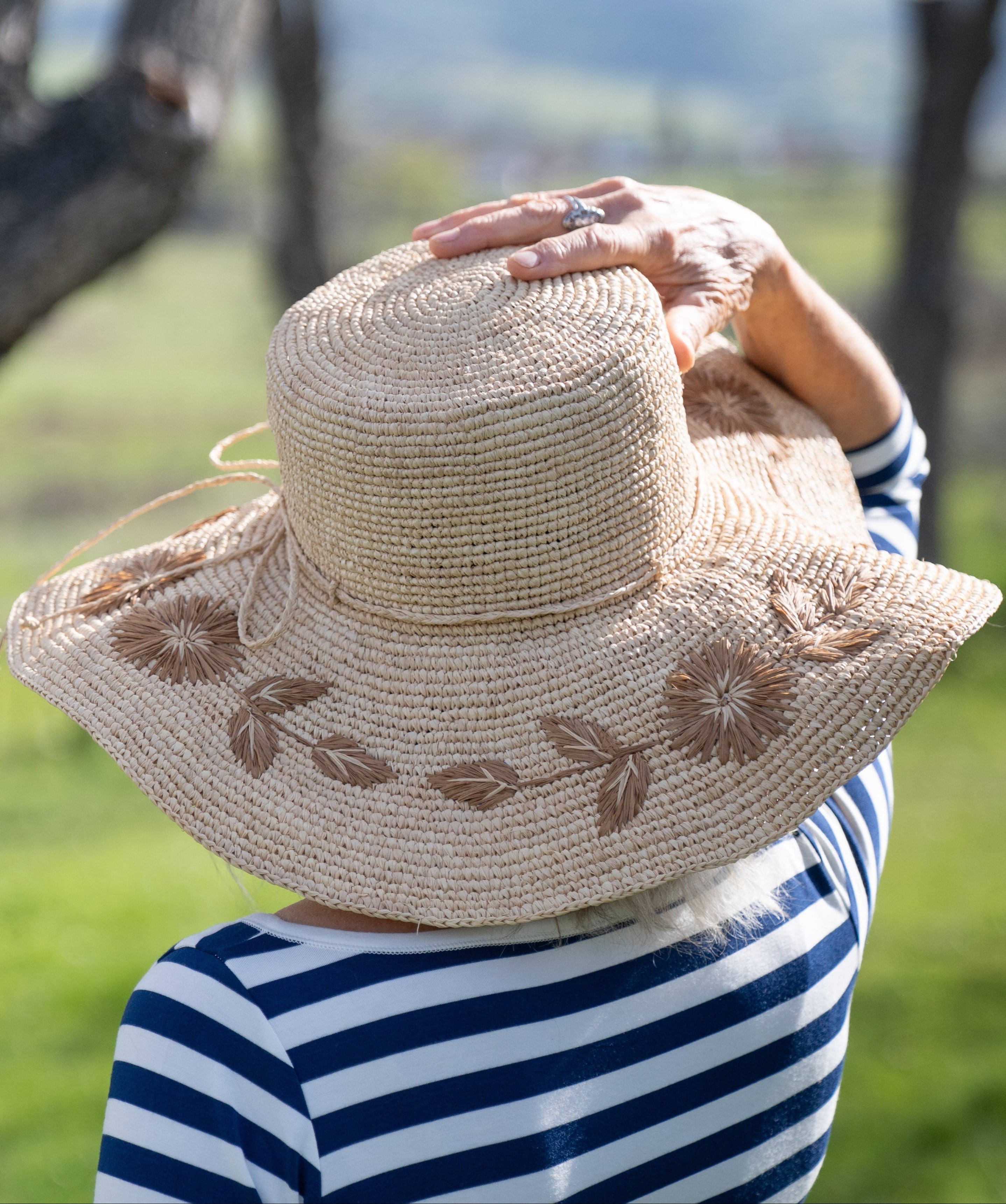 Model wearing 5.5" Brim Gigi Natural and Cinnamon Floral Embroidered Crochet Straw Sun Hats handmade crochet natural raffia palm fiber in a solid hue of natural straw color making a concentric pattern from the center top of the flattened crown spiraling out to the edge of the brim with matching adjustable raffia braid hat band and cinnamon dark brown floral pattern with leaves embroidered on the brim - Shebobo