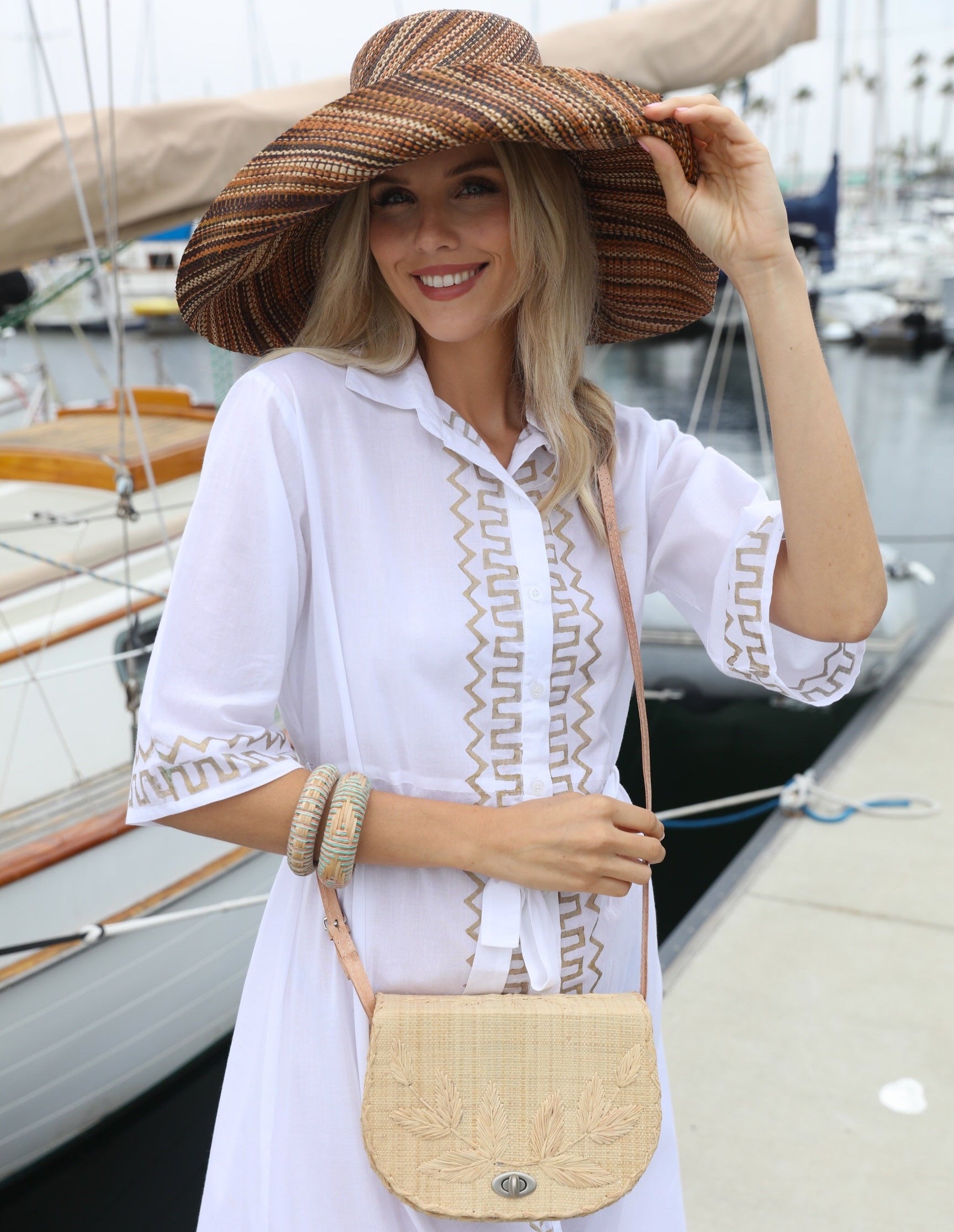 Model wearing 70's Crossbody Straw Bag handmade loomed natural raffia palm fiber purse with natural colored accent cross stitch edging and matching leaf design embroidery handbag with adjustable leather strap - Shebobo (with Monica 7" Wide Brim Cinnamon/Tobacco/Brown Multicolor Melange Packable Straw Sun Hat)