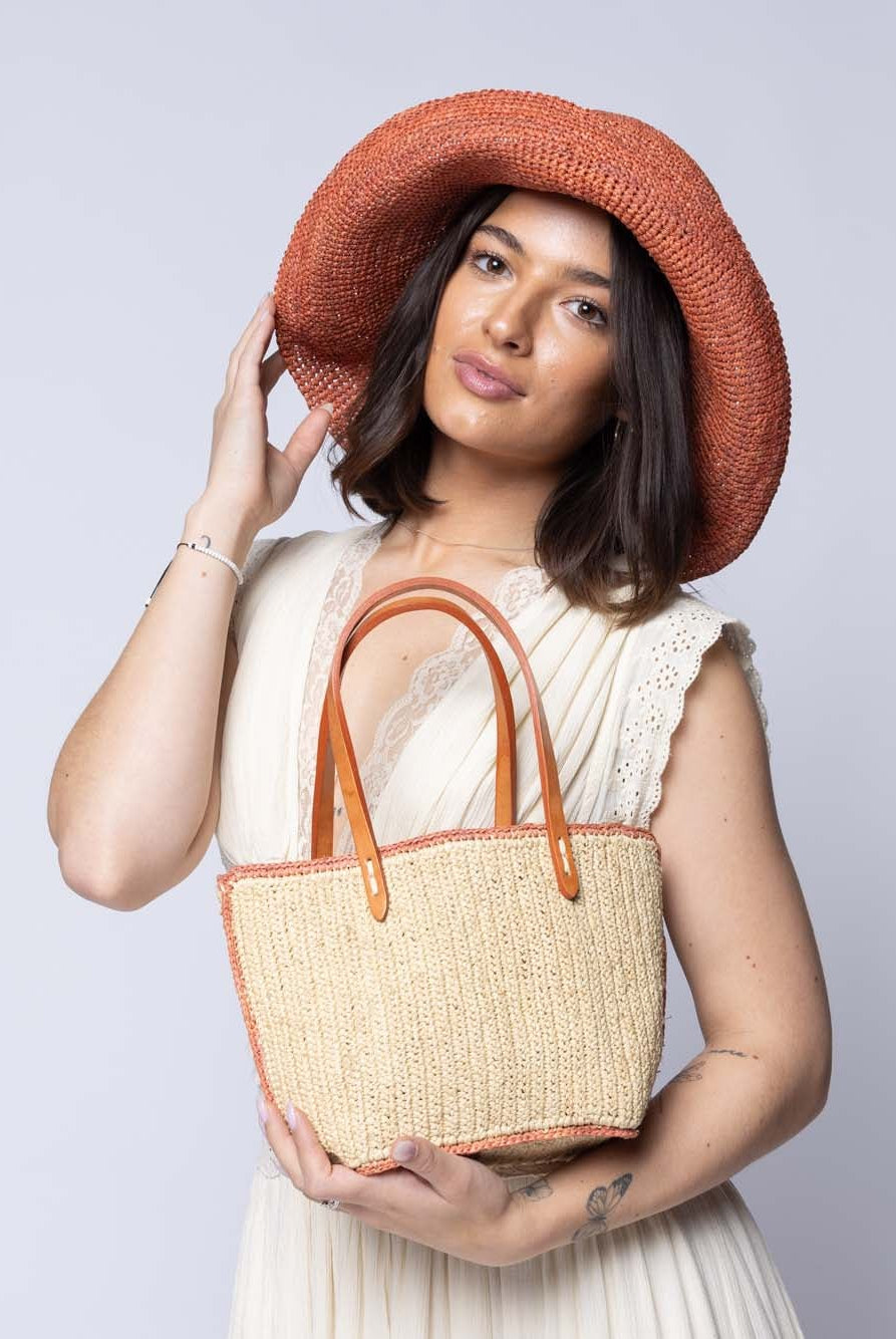 Model wearing Side view Adele Mini Crochet Raffia Straw Handbag Purse with Colored Edge Natural & Blush handmade crochet raffia palm fiber in a solid hue of natural straw color with contrasting narrow band of blush orange/pink around the outer edges of the bag with subtle vertical band texture and leather handles - Shebobo