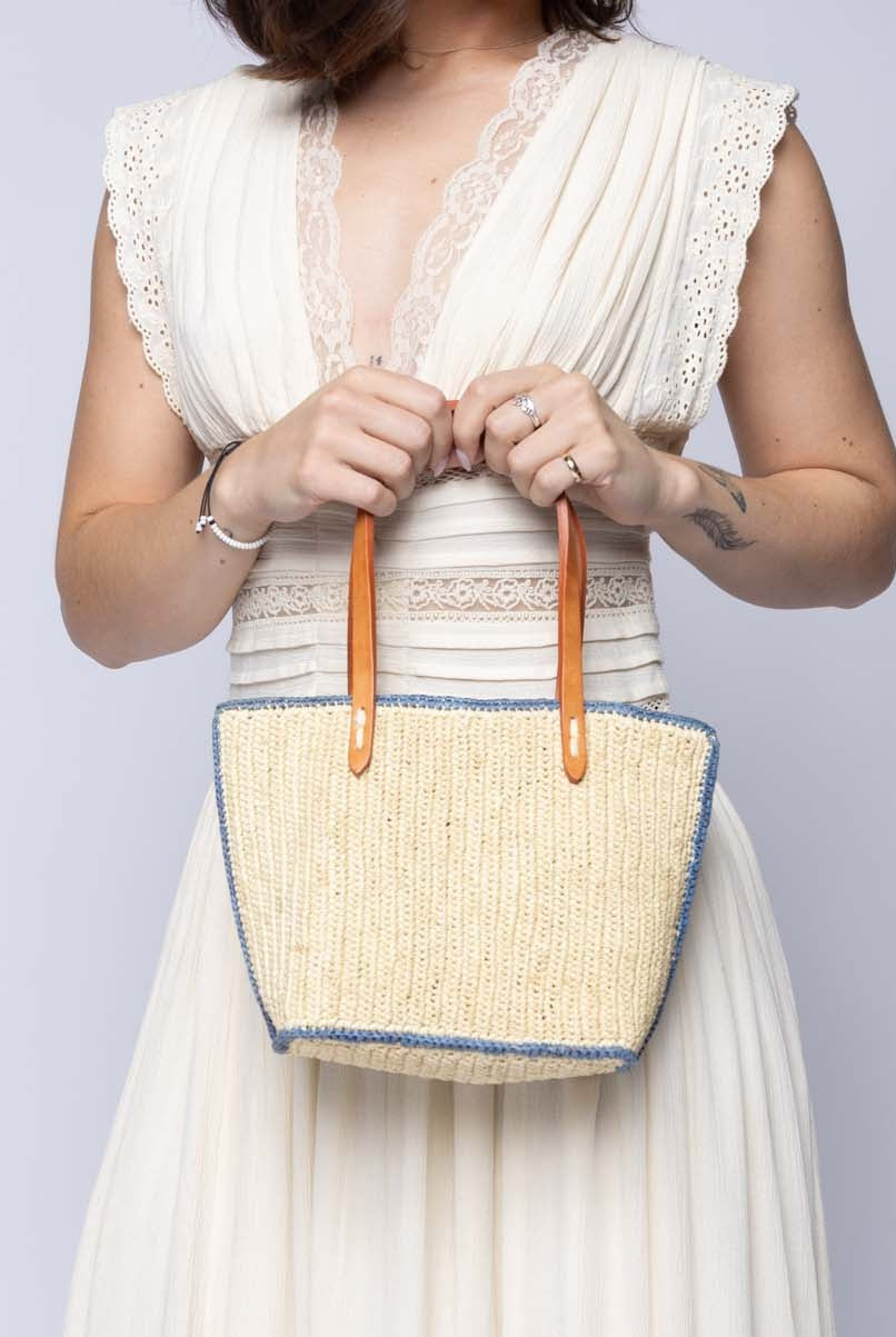 Model wearing Adele Mini Crochet Raffia Straw Handbag Purse with Colored Edge Natural & Denim handmade crochet raffia palm fiber in a solid hue of natural straw color with contrasting narrow band of denim light/blue around the outer edges of the bag with subtle vertical band texture and leather handles - Shebobo
