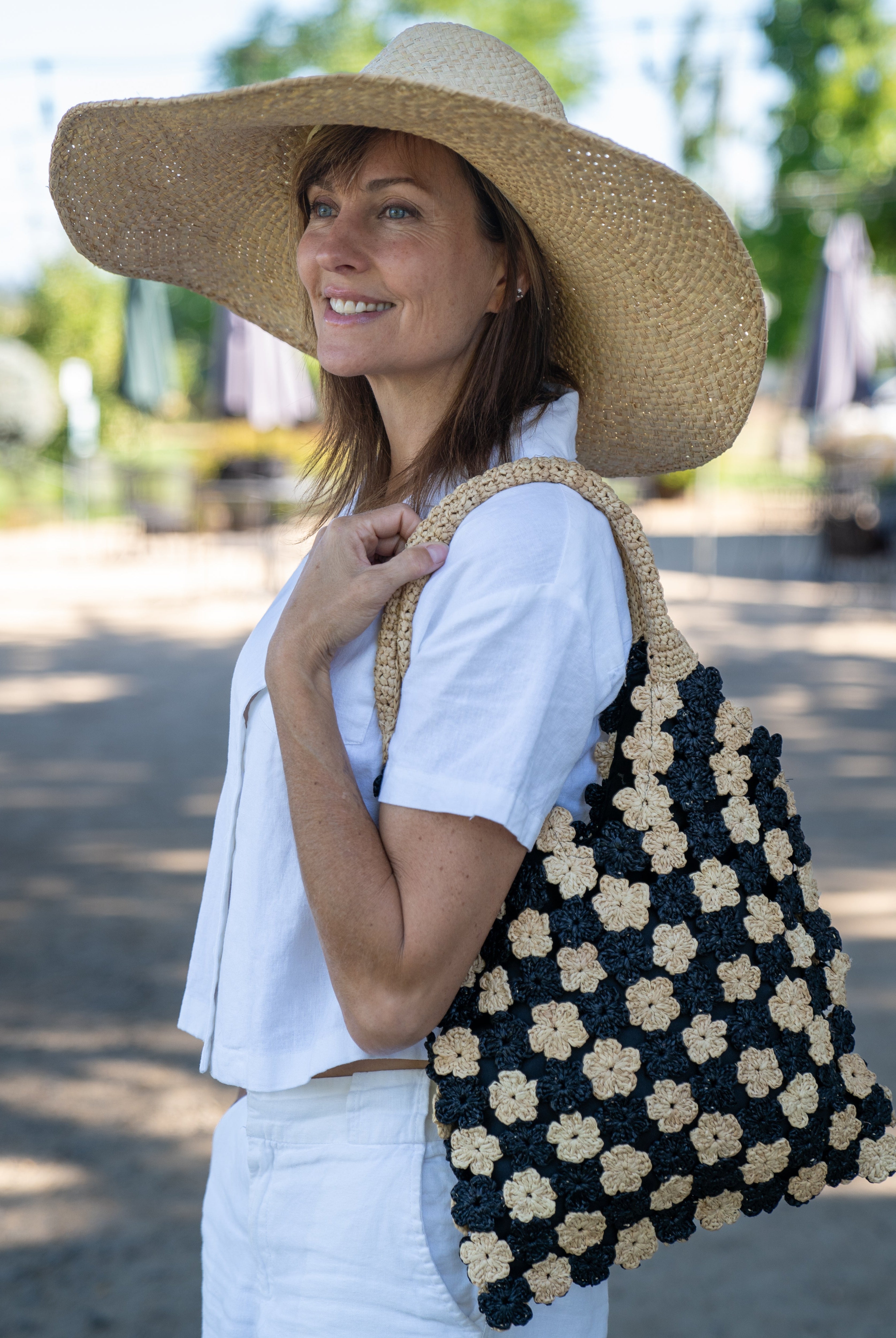 Model wearing Amalie Two Tone Daisy Flower Crochet Straw Handbag handmade crochet mini flower buds from crochet natural raffia palm fiber in an alternating colored pattern of black and natural straw color with black internal liner and natural woven handles purse - Shebobo (with Natasha 7" Wide Brim Woven XL Straw Sun Hats)