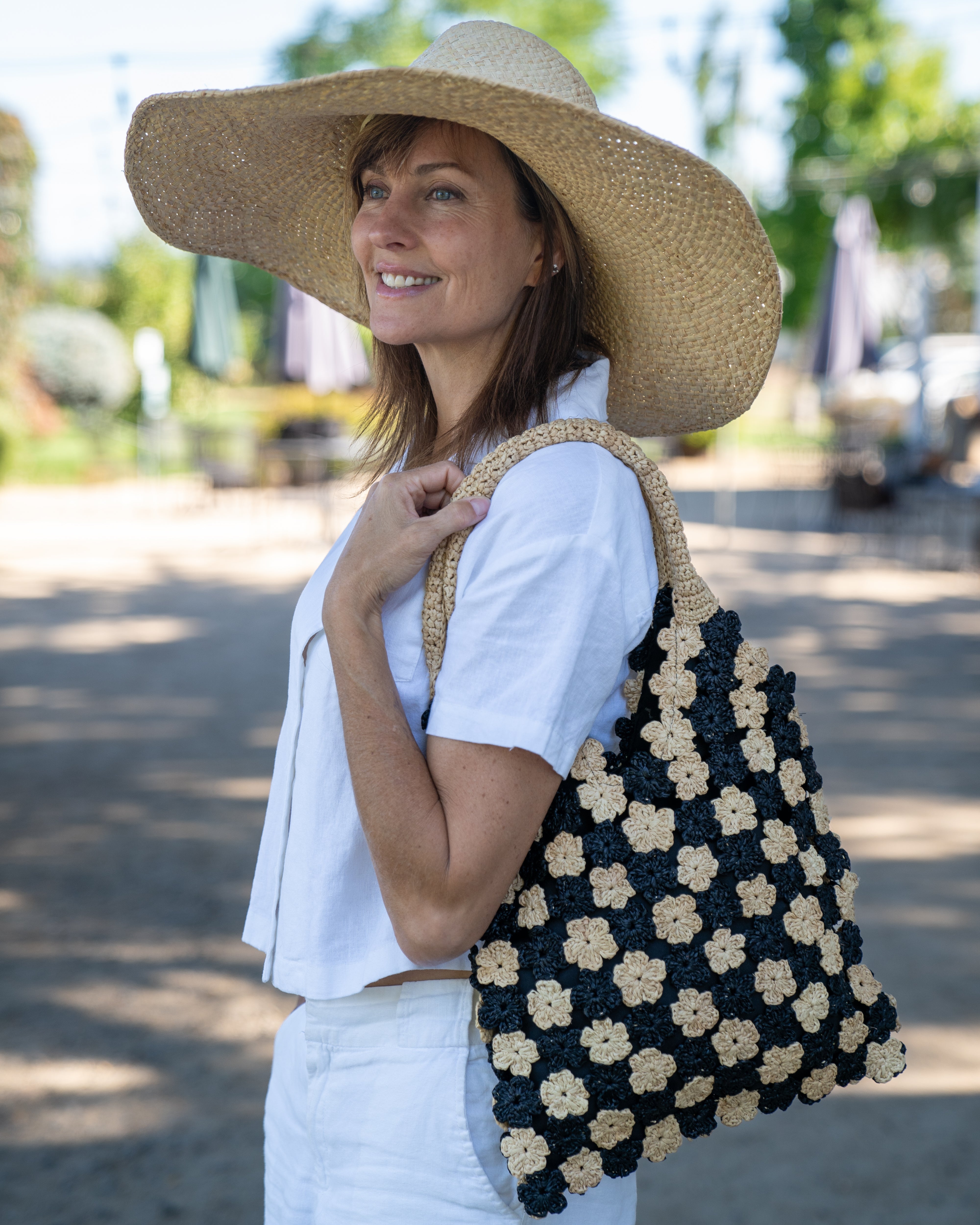 Model wearing Amalie Two Tone Daisy Flower Crochet Straw Handbag handmade crochet mini flower buds from crochet natural raffia palm fiber in an alternating colored pattern of black and natural straw color with black internal liner and natural woven handles purse - Shebobo (with Natasha 7" Wide Brim Woven XL Straw Sun Hats)