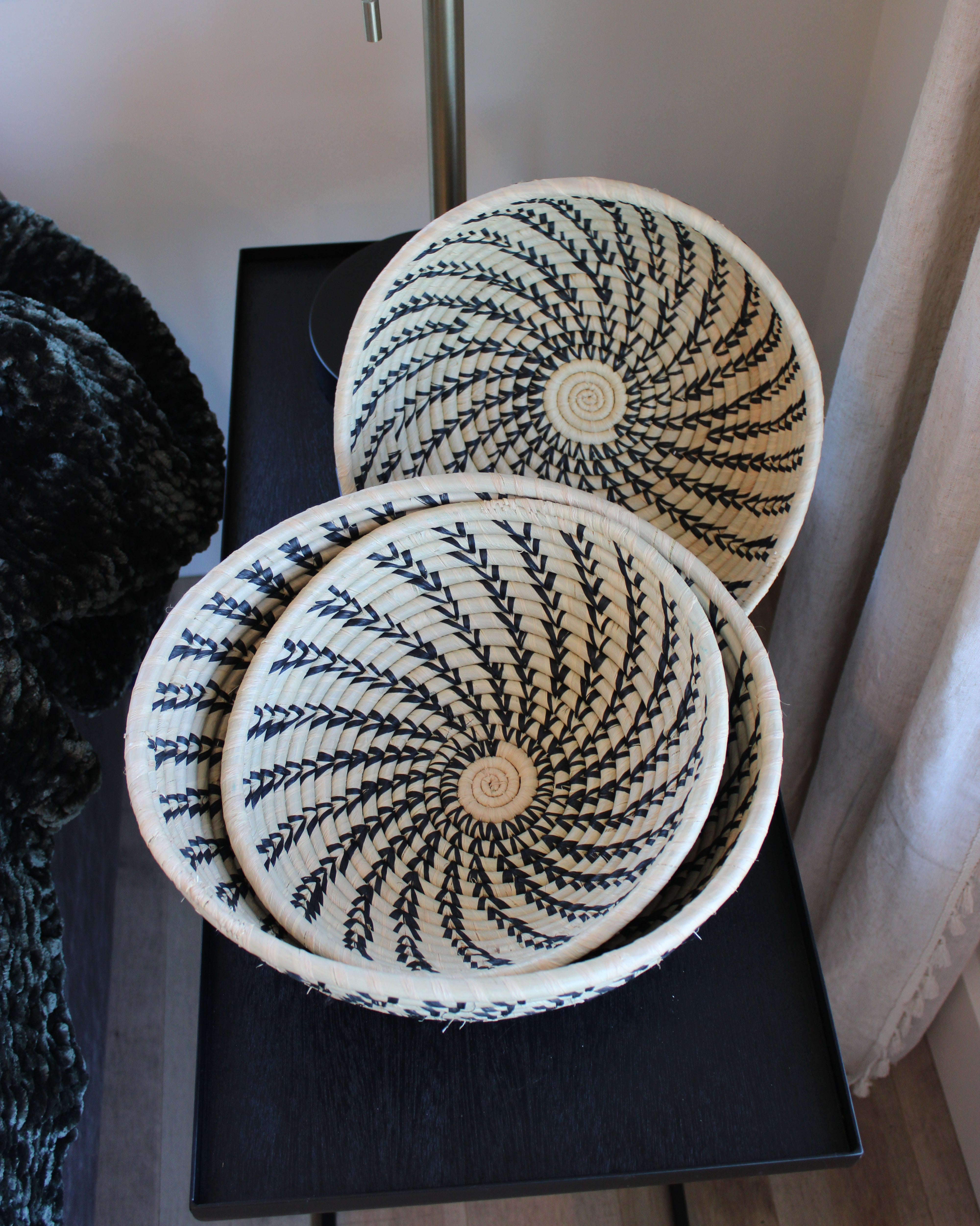 Home decor lifestyle view Amara Raffia Straw Decor Display Basket Bowls: Set of 3 handmade woven natural raffia palm fiber in a crochet loop technique concentric coiled circular pattern with natural straw color centered in the bottom and that go up the sides of the bowls with contrasting black swirl stitch pattern and matching natural rim/edge set of small, medium, and large straw display bowls - Shebobo
