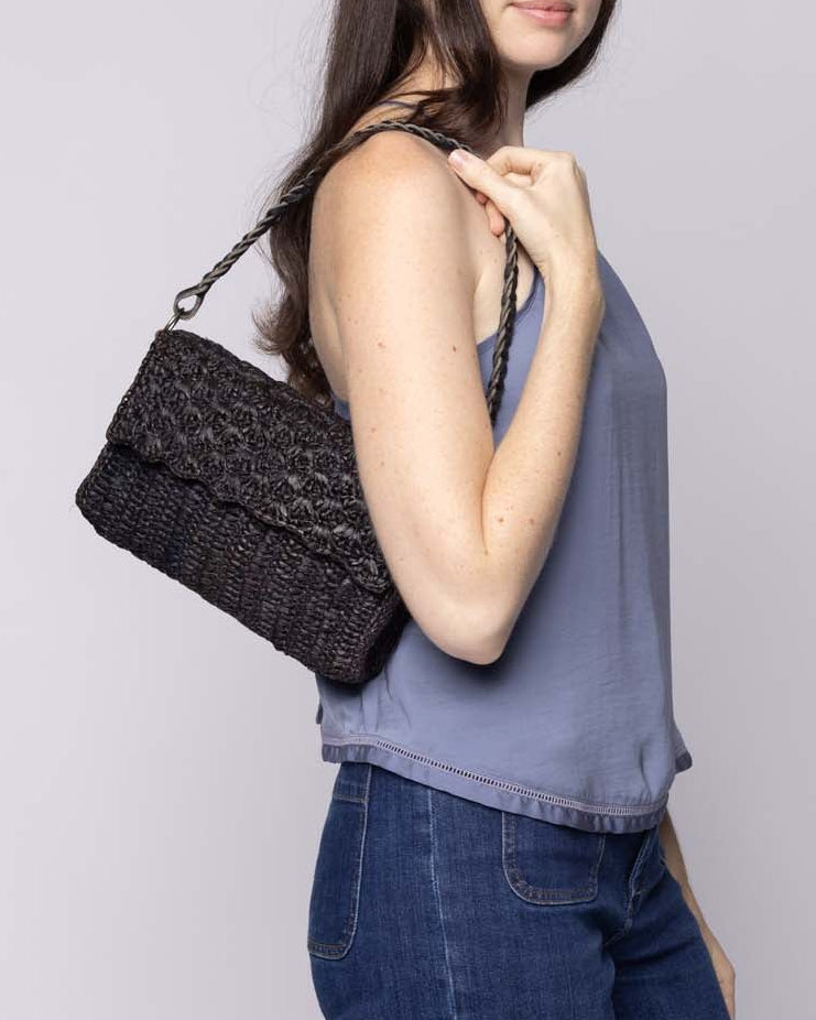 Model wearing Anna Petal Pattern Crochet Straw Over Shoulder Envelope Clutch Purse handmade raffia palm fiber in a solid hue of Black crochet in multiple textures resulting in a subtle pattern of vertical linked bands on the bottom half of the bag, and a floral petal or shell pattern on the upper envelope flap with braided leather shoulder strap handbag shoulder bag - Shebobo