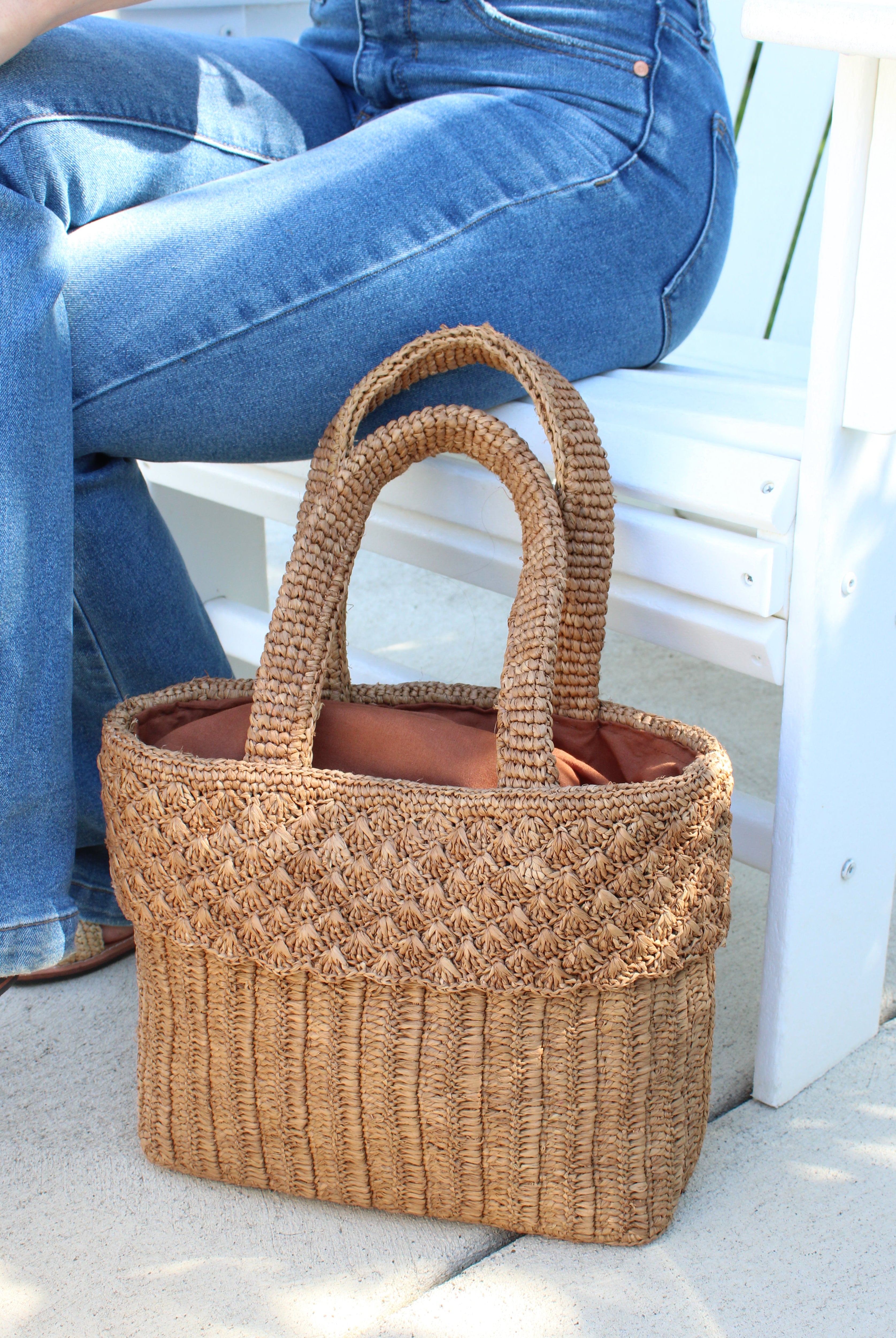 Model wearing Anna Petal Pattern Crochet Straw Basket Handbag handmade natural raffia palm fiber in a solid hue of Cappuccino tea/light/brown crochet in multiple textures resulting in a subtle pattern of vertical linked bands on the bottom 2/3 of the bag, and a floral petal or shell pattern on the upper third with rolled panel of links making up the handles Beach Bag Purse - Shebobo