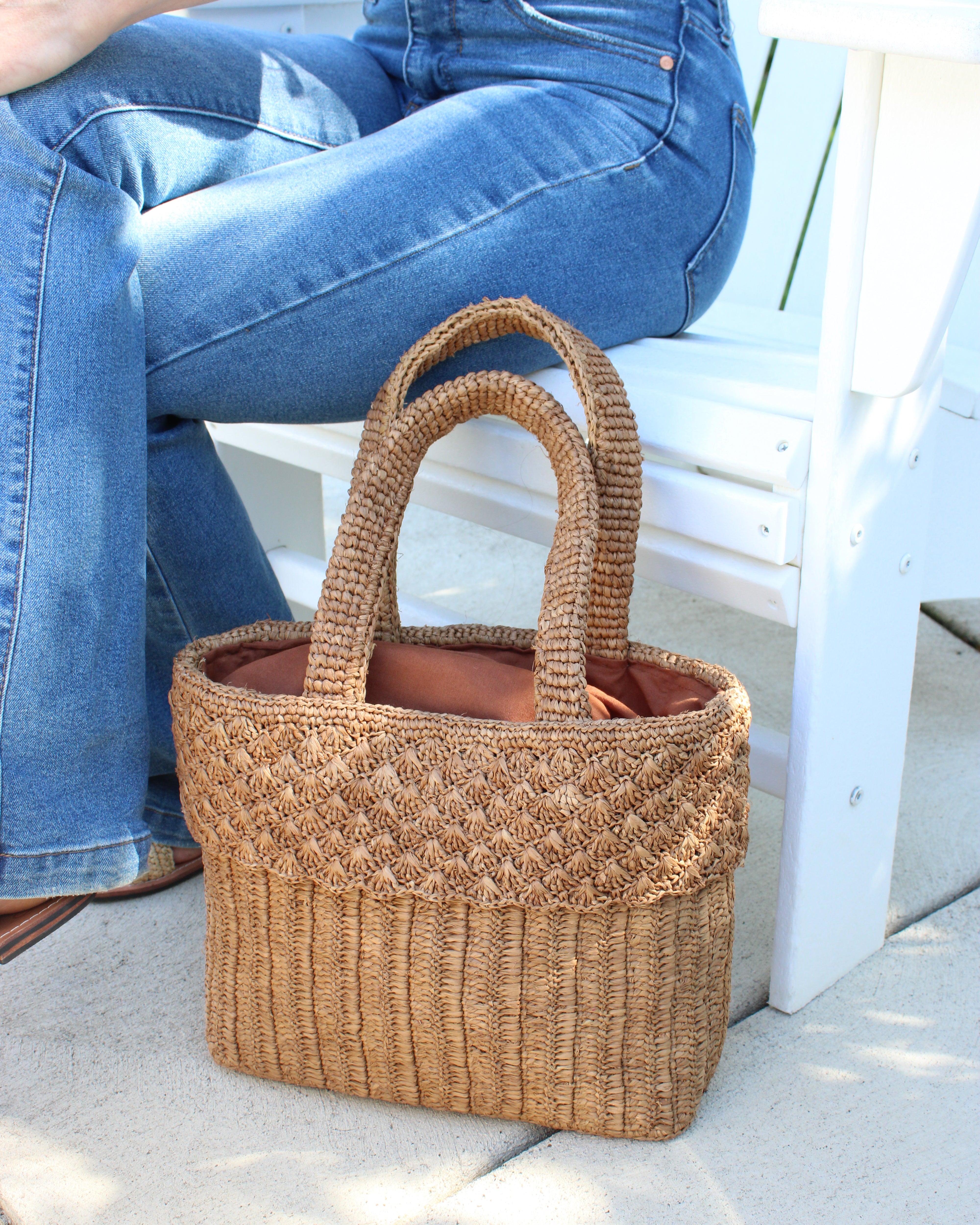 Model wearing Anna Petal Pattern Crochet Straw Basket Handbag handmade natural raffia palm fiber in a solid hue of Cappuccino tea/light/brown crochet in multiple textures resulting in a subtle pattern of vertical linked bands on the bottom 2/3 of the bag, and a floral petal or shell pattern on the upper third with rolled panel of links making up the handles Beach Bag Purse - Shebobo