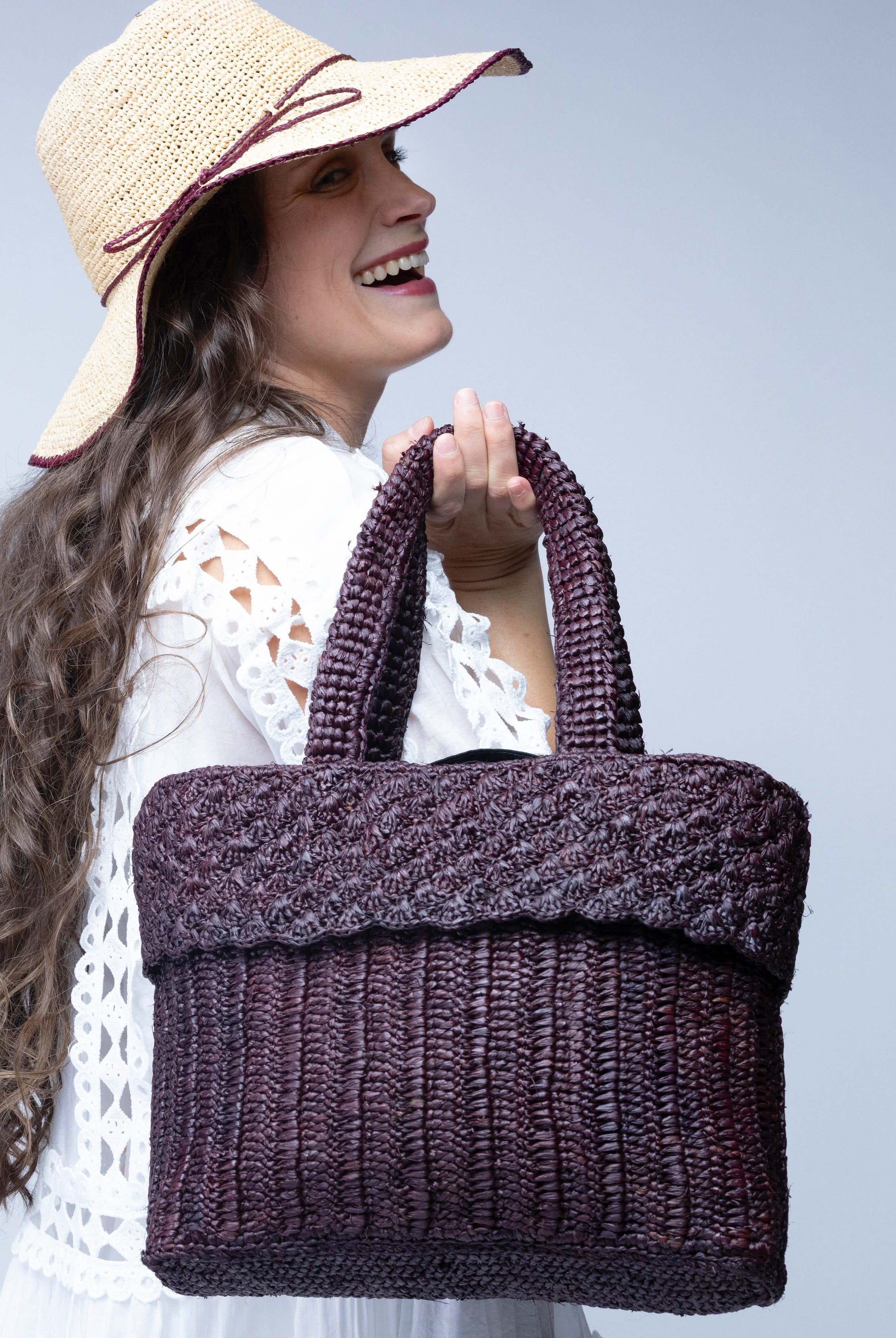 Model wearing Anna Petal Pattern Crochet Straw Basket Handbag handmade natural raffia palm fiber in a solid hue of Espresso chocolate/dark/brown crochet in multiple textures resulting in a subtle pattern of vertical linked bands on the bottom 2/3 of the bag, and a floral petal or shell pattern on the upper third with rolled panel of links making up the handles Beach Bag Purse - Shebobo (with Rachel 4" Brim Crochet Straw Sun Hats with Colored Edge)