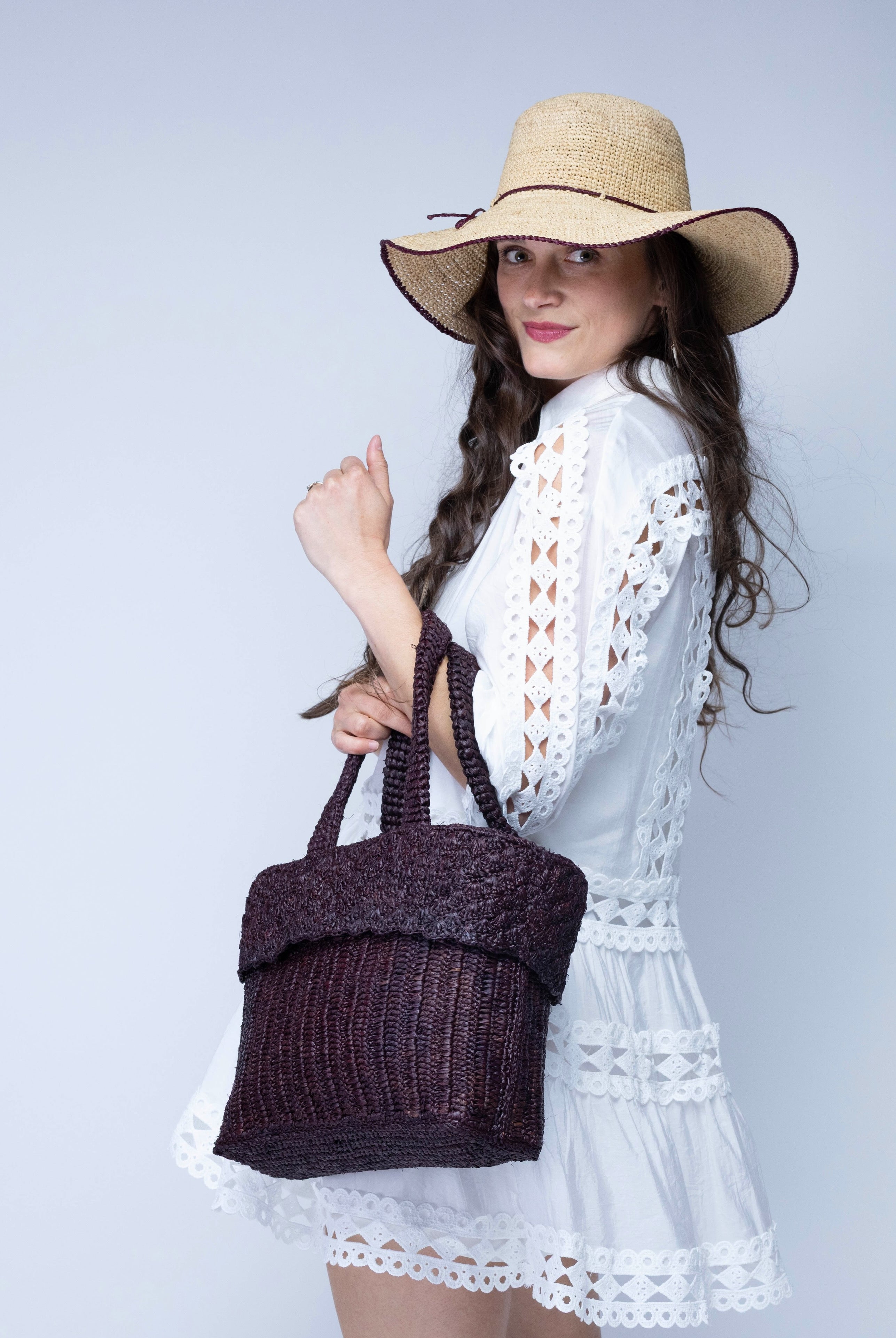 Model wearing Anna Petal Pattern Crochet Straw Basket Handbag handmade natural raffia palm fiber in a solid hue of Mocha chocolate/dark/brown crochet in multiple textures resulting in a subtle pattern of vertical linked bands on the bottom 2/3 of the bag, and a floral petal or shell pattern on the upper third with rolled panel of links making up the handles Beach Bag Purse - Shebobo (with Rachel 4" Brim Crochet Straw Sun Hats with Colored Edge)