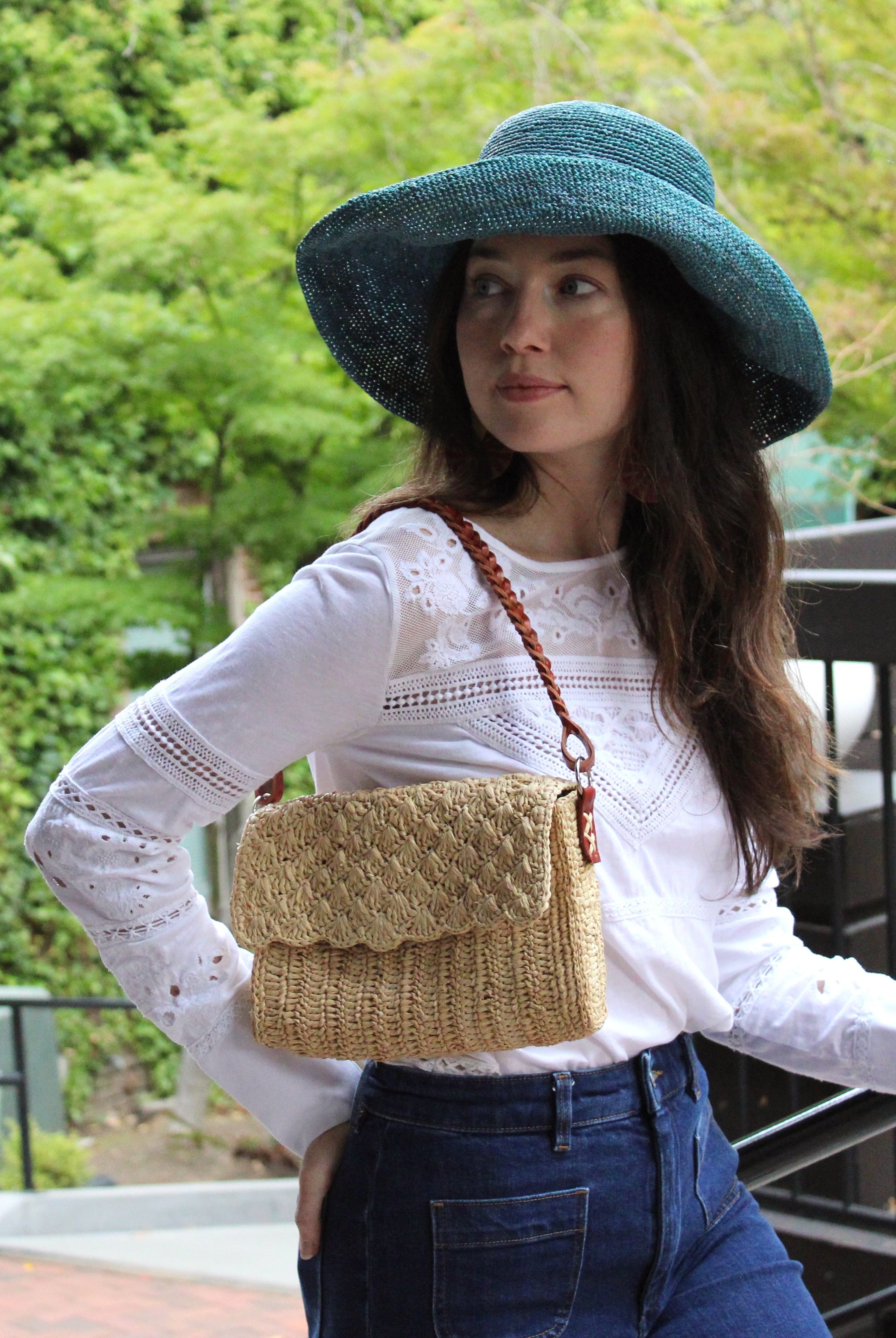 Model wearing Anna Petal Pattern Crochet Straw Over Shoulder Envelope Clutch Purse handmade raffia palm fiber in a solid hue of Natural straw color crochet in multiple textures resulting in a subtle pattern of vertical linked bands on the bottom half of the bag, and a floral petal or shell pattern on the upper envelope flap with braided leather shoulder strap handbag shoulder bag - Shebobo (with Leor 3" Brim Crochet Straw Sun Hats)