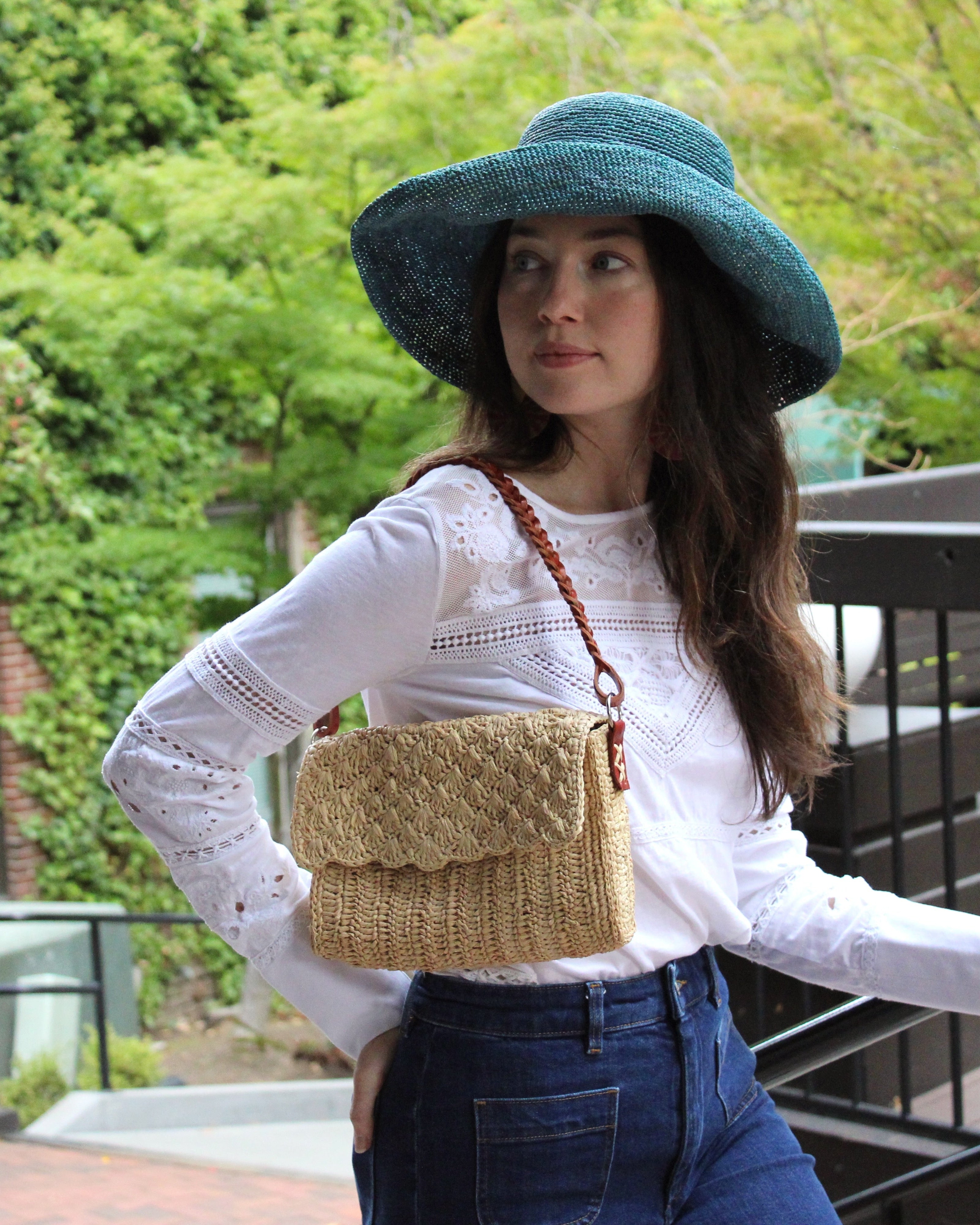 Model wearing Anna Petal Pattern Crochet Straw Over Shoulder Envelope Clutch Purse handmade raffia palm fiber in a solid hue of Natural straw color crochet in multiple textures resulting in a subtle pattern of vertical linked bands on the bottom half of the bag, and a floral petal or shell pattern on the upper envelope flap with braided leather shoulder strap handbag shoulder bag - Shebobo (with Leor 3" Brim Crochet Straw Sun Hats)