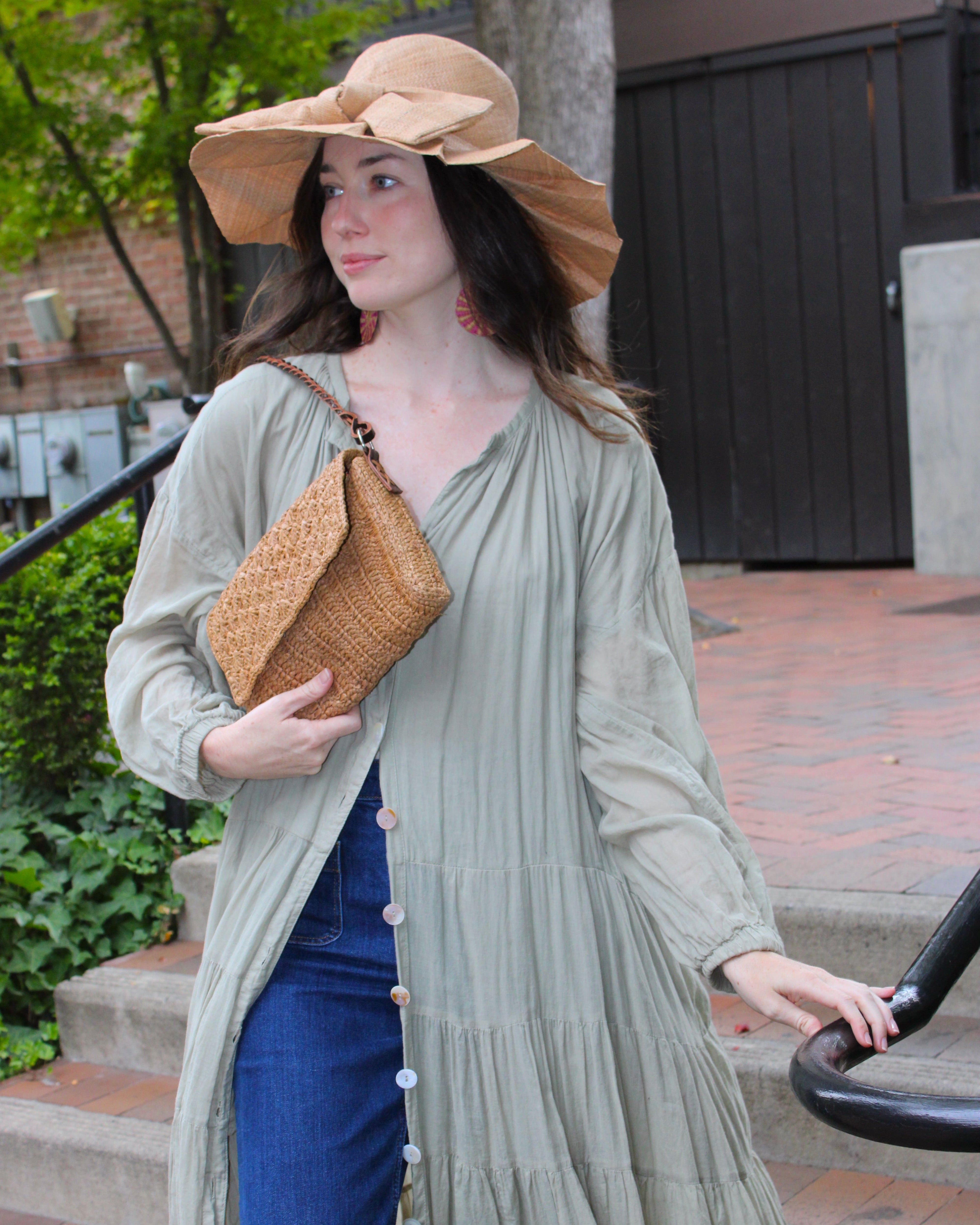 Model wearing Anna Petal Pattern Crochet Straw Over Shoulder Envelope Clutch Purse handmade raffia palm fiber in a solid hue of Cappuccino tea/light/brown crochet in multiple textures resulting in a subtle pattern of vertical linked bands on the bottom half of the bag, and a floral petal or shell pattern on the upper envelope flap with braided leather shoulder strap handbag shoulder bag - Shebobo (with Lilly 5" Brim Fan Pleated Straw Sun Hats with Big Bow)
