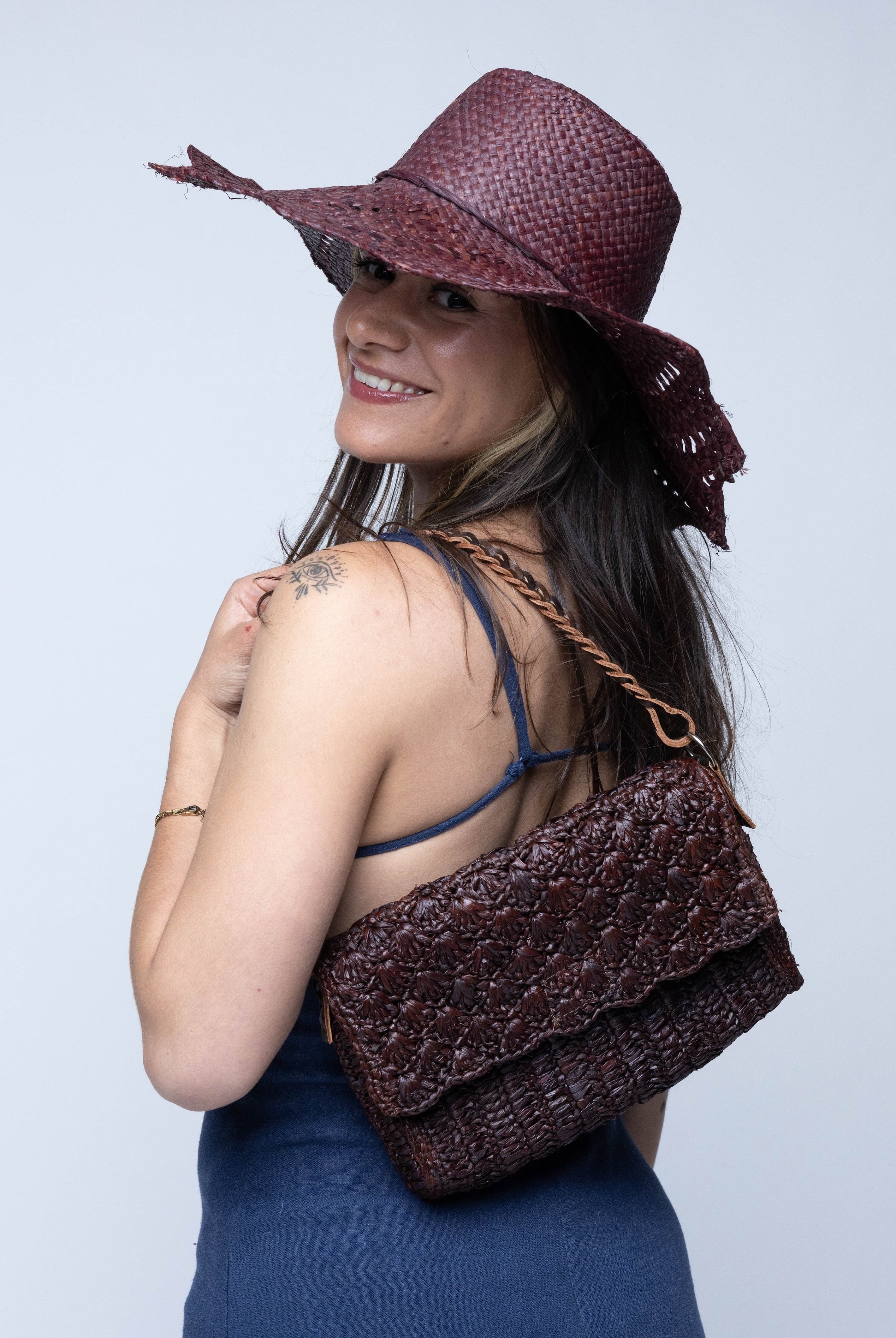 Model wearing Anna Petal Pattern Crochet Straw Over Shoulder Envelope Clutch Purse handmade raffia palm fiber in a solid hue of Mocha chocolate/dark/brown crochet in multiple textures resulting in a subtle pattern of vertical linked bands on the bottom half of the bag, and a floral petal or shell pattern on the upper envelope flap with braided leather shoulder strap handbag shoulder bag - Shebobo (with Sara 4.5" Brim Woven Straw Sun Hats with Zigzag Edge)