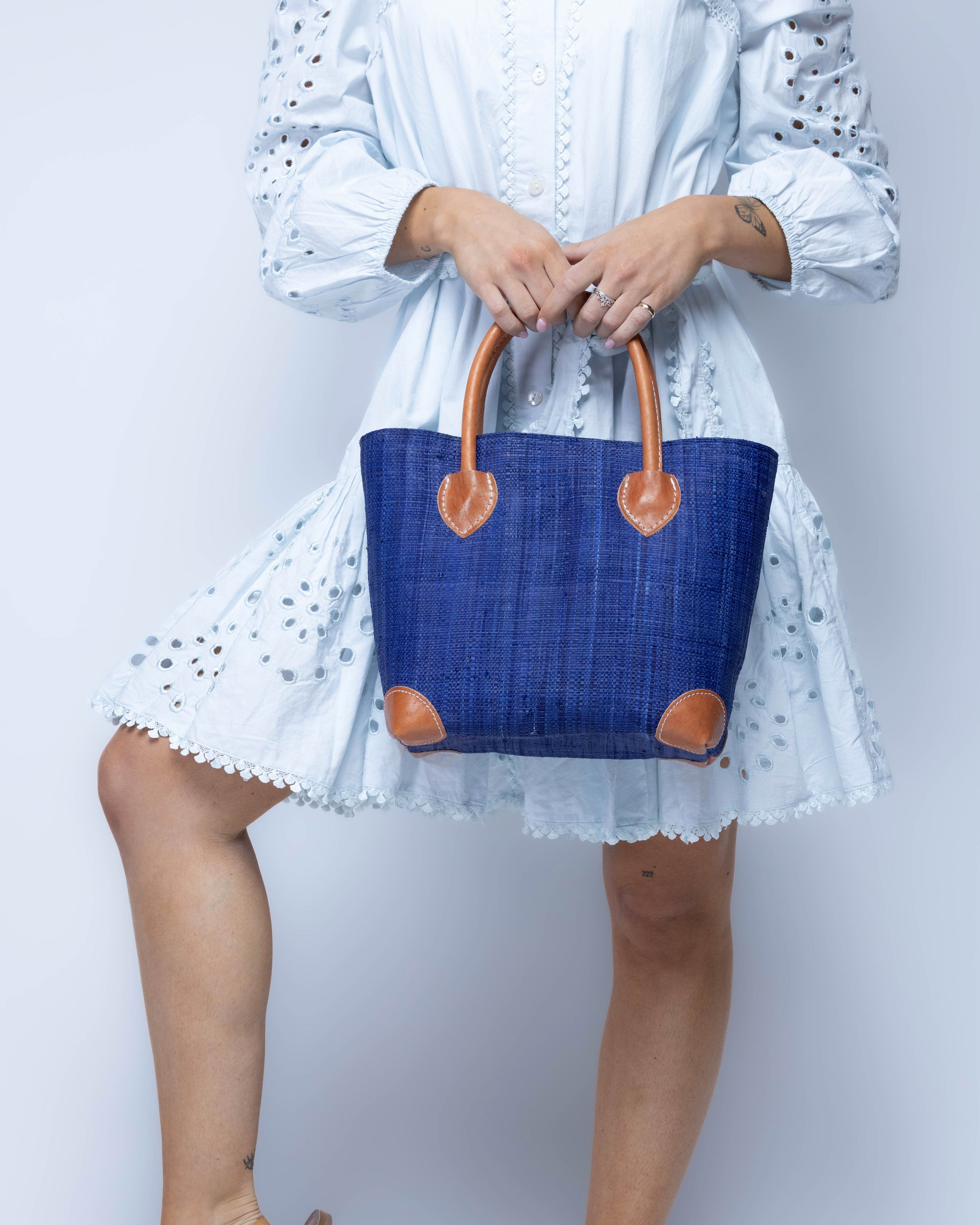 Model wearing Small Augustine Straw Basket Bag Azure handmade loomed raffia palm fiber in a solid hue of azure dusk/navy/dark/blue handbag purse with leather accents and handles beach bag - Shebobo