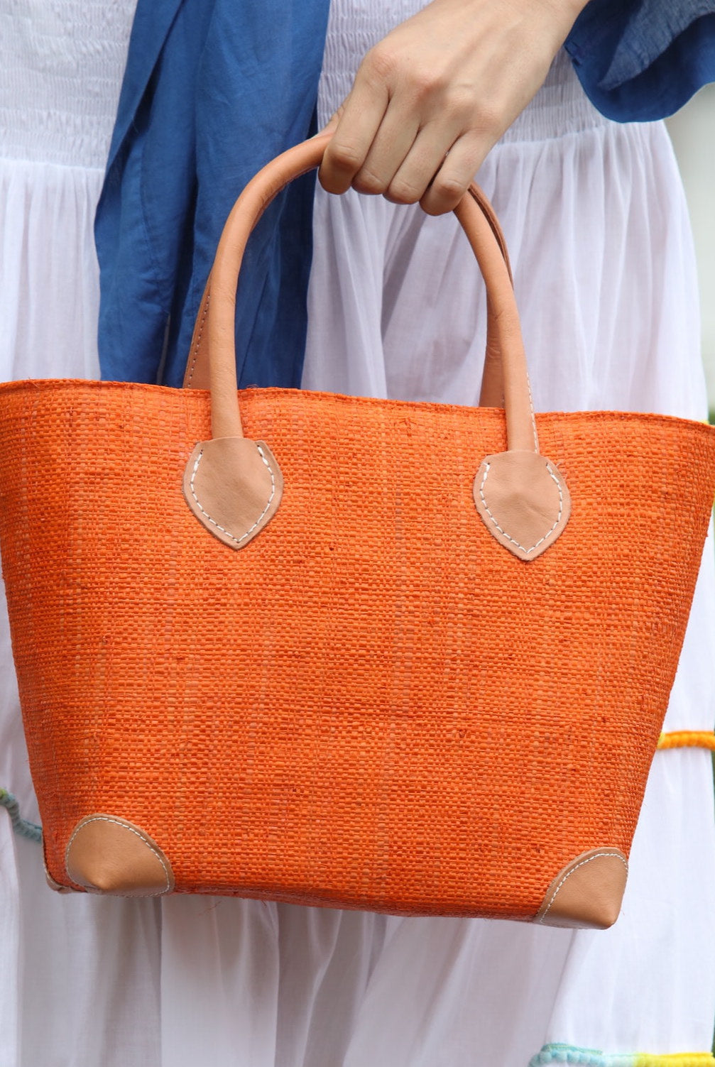 Model wearing Augustine Straw Basket Bag handmade loomed raffia handbag with leather corners and handles in coral orange/red - Shebobo