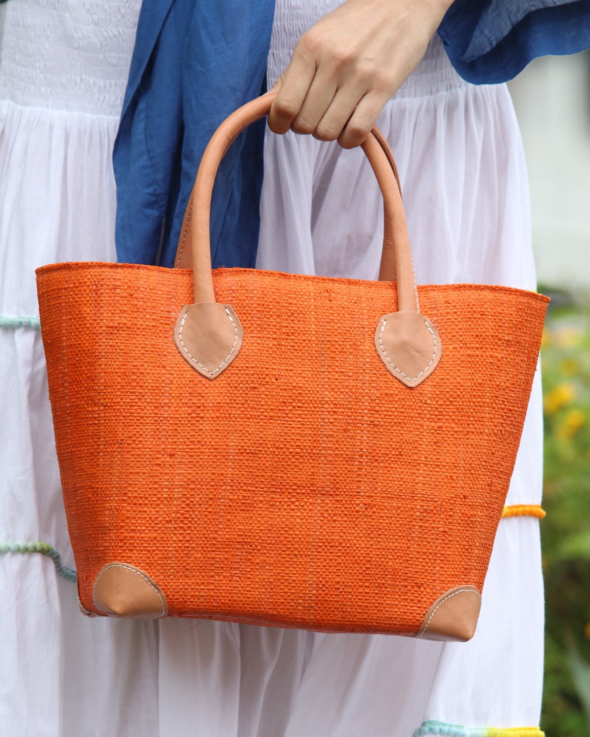 Model wearing Augustine Straw Basket Bag handmade loomed raffia handbag with leather corners and handles in coral orange/red - Shebobo