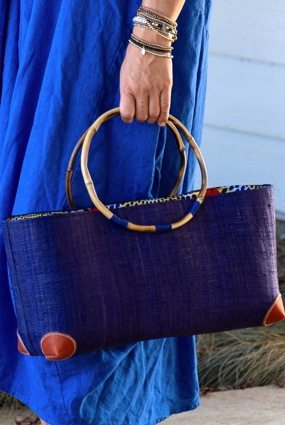 Model wearing Bebe Straw Handbag with Bamboo Handle handmade loomed natural raffia palm fiber in a solid hue of Dusk dark/navy/blue with molded circular bamboo handles, leather feet, and assorted print African wax-cloth fabric liner - Shebobo