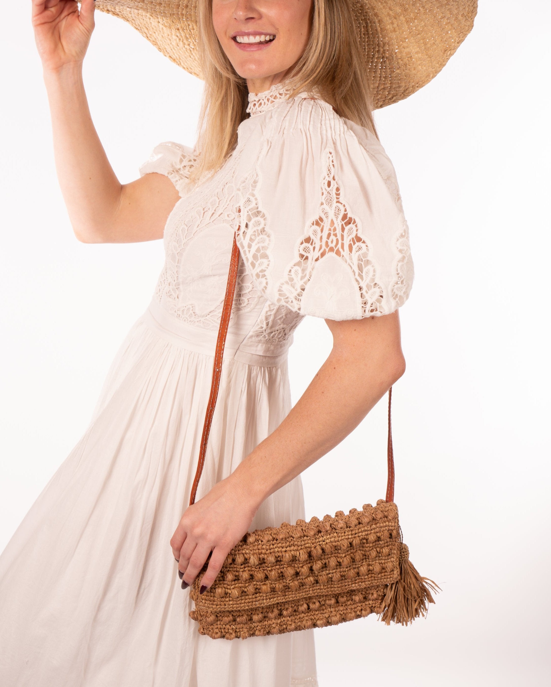 Model wearing Boucle Cappuccino Crochet Straw Crossbody Bag with Tassel Charm handmade woven raffia palm fiber in a solid hue of cappuccino light/brown/beige in a supple nub/ball texture of horizontal bands around the bag with link weave around the outer envelope closure flap plus adjustable leather shoulder strap beach bag purse - Shebobo (with Vanessa Wide Brim Woven Straw Sun Hat)