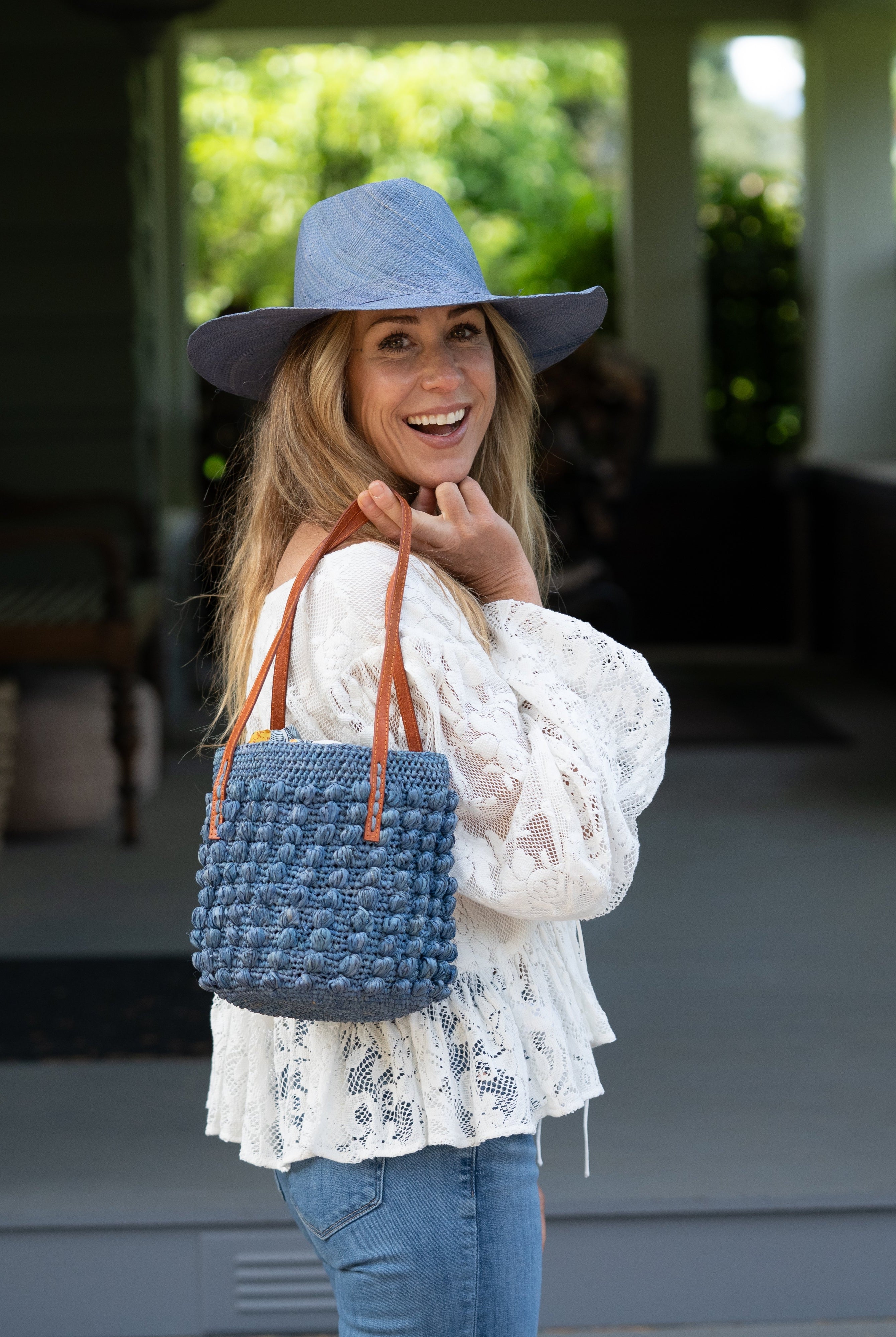 Model wearing Boucle Denim Crochet Small Straw Handbag handmade woven raffia palm fiber in a solid hue of denim retro/light/blue in a supple nub/ball texture of horizontal bands around the bag with link weave around the top plus assorted print drawstring liner and leather handles shoulder bag purse - Shebobo (with Panama Unisex Straw Hats)