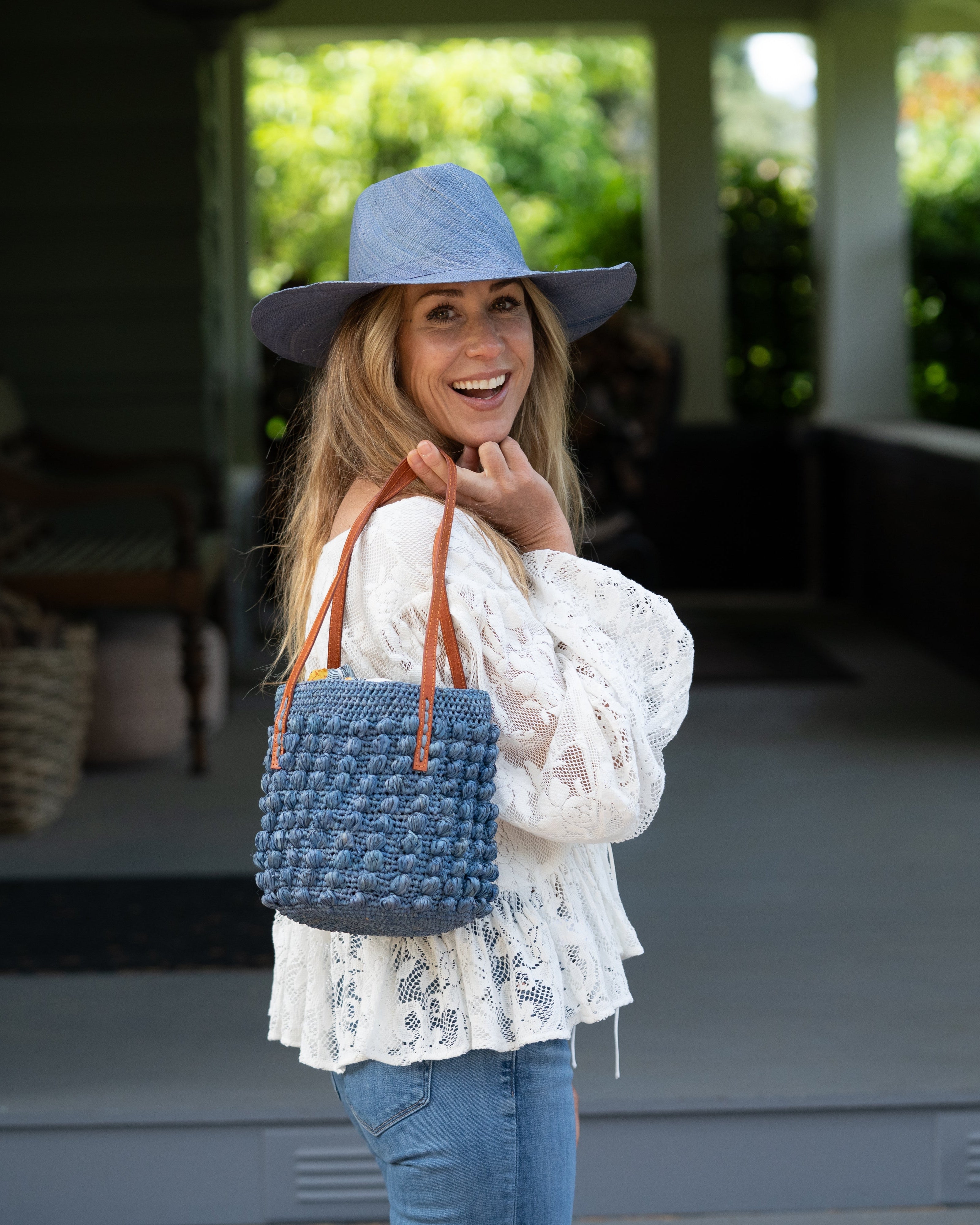 Model wearing Boucle Denim Crochet Small Straw Handbag handmade woven raffia palm fiber in a solid hue of denim retro/light/blue in a supple nub/ball texture of horizontal bands around the bag with link weave around the top plus assorted print drawstring liner and leather handles shoulder bag purse - Shebobo (with Panama Unisex Straw Hats)