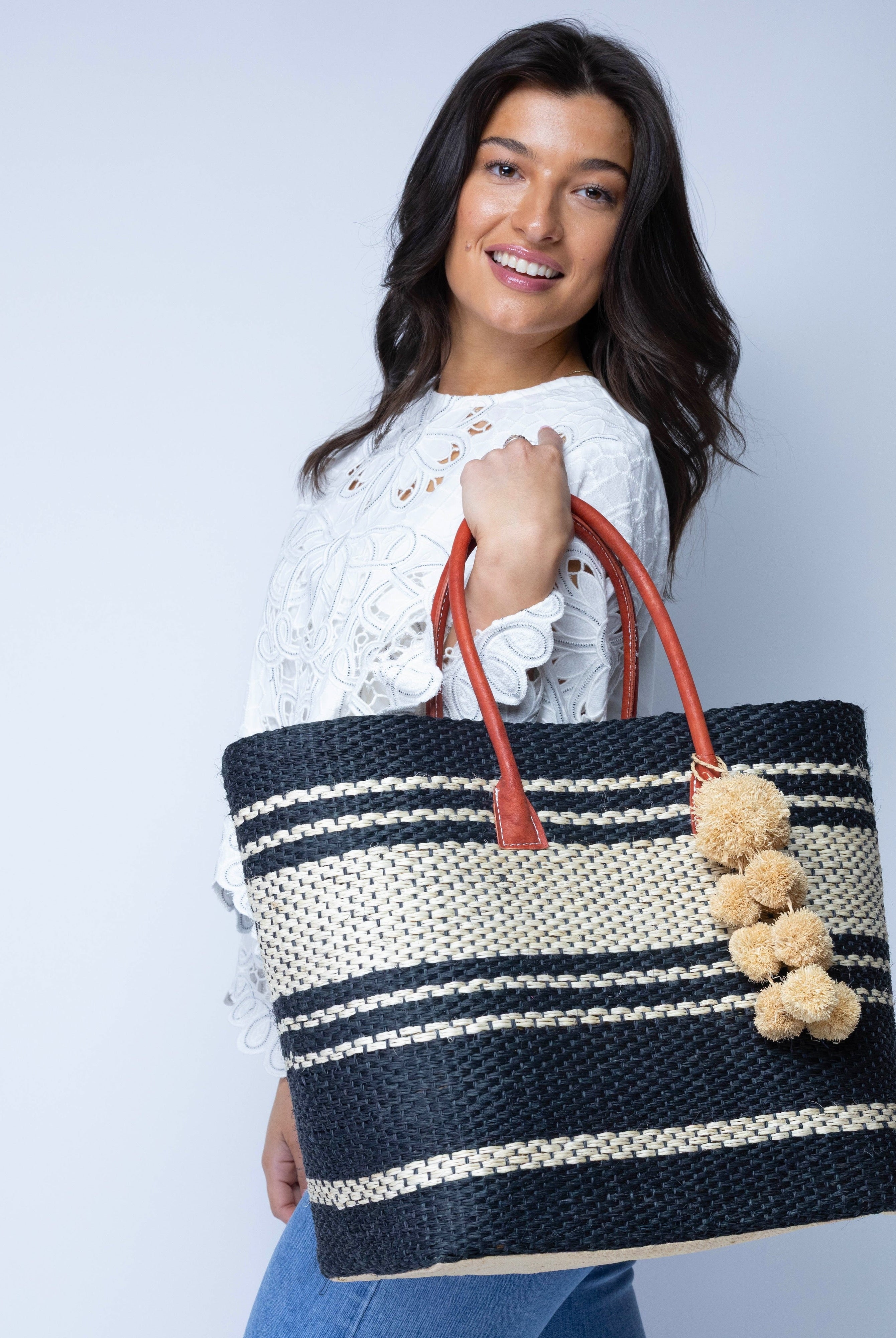 Model wearing Cabrillo Sisal Basket Bag with Waterfall Pompoms Charm Embellishment handmade natural sisal fiber woven into a multicolor two tone Black and natural straw color horizontal stripe pattern with muti-width bands of color plus leather handles and natural raffia charm shoulder bag beach bag - Shebobo