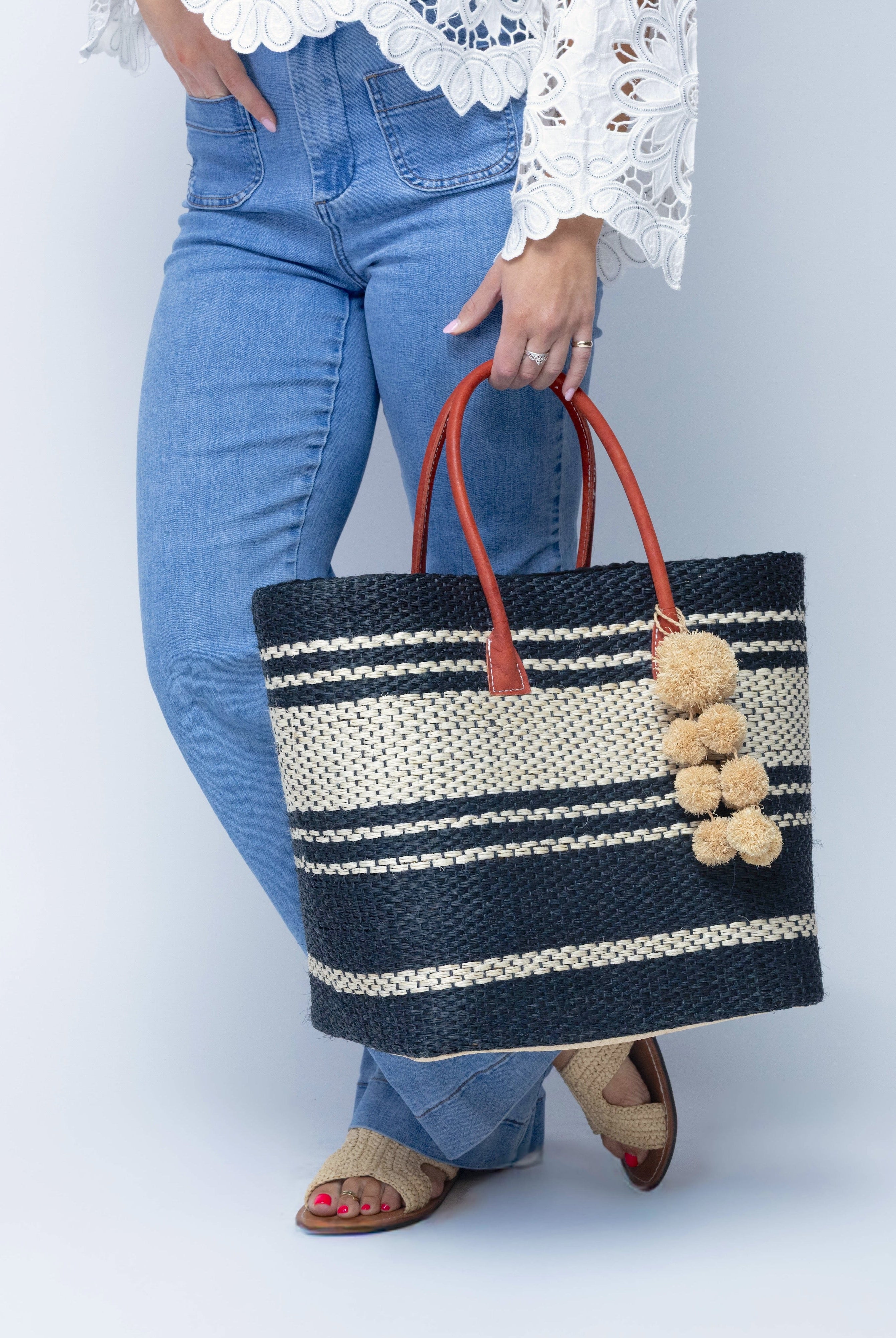 Model wearing Cabrillo Sisal Basket Bag with Waterfall Pompoms Charm Embellishment handmade natural sisal fiber woven into a multicolor two tone Black and natural straw color horizontal stripe pattern with muti-width bands of color plus leather handles and natural raffia charm shoulder bag beach bag - Shebobo