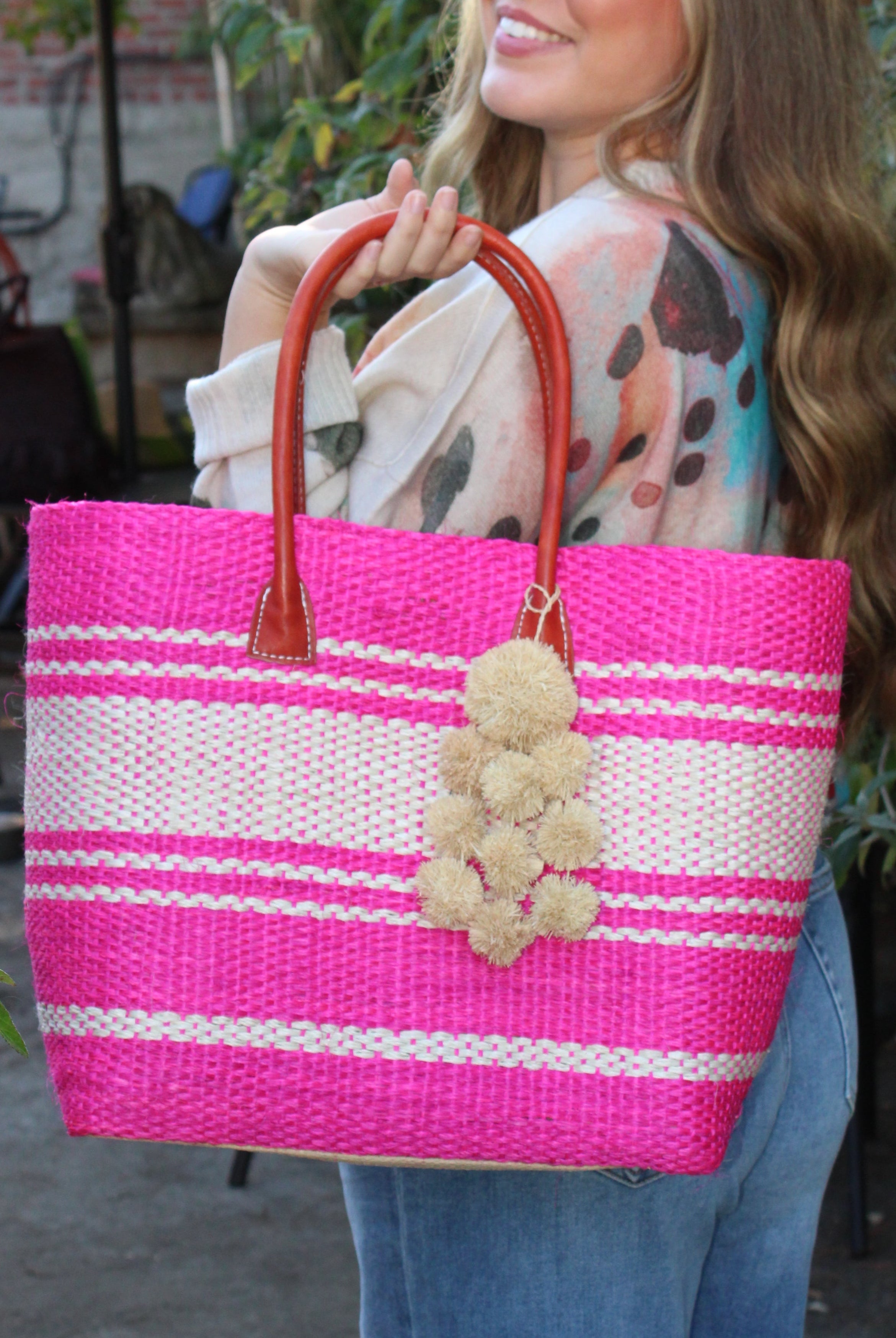 Model wearing Cabrillo Sisal Basket Bag with Waterfall Pompoms Charm Embellishment handmade natural sisal fiber woven into a multicolor two tone Fuchsia hot/bright/barbie/pink and natural straw color horizontal stripe pattern with muti-width bands of color plus leather handles and natural raffia charm shoulder bag beach bag - Shebobo
