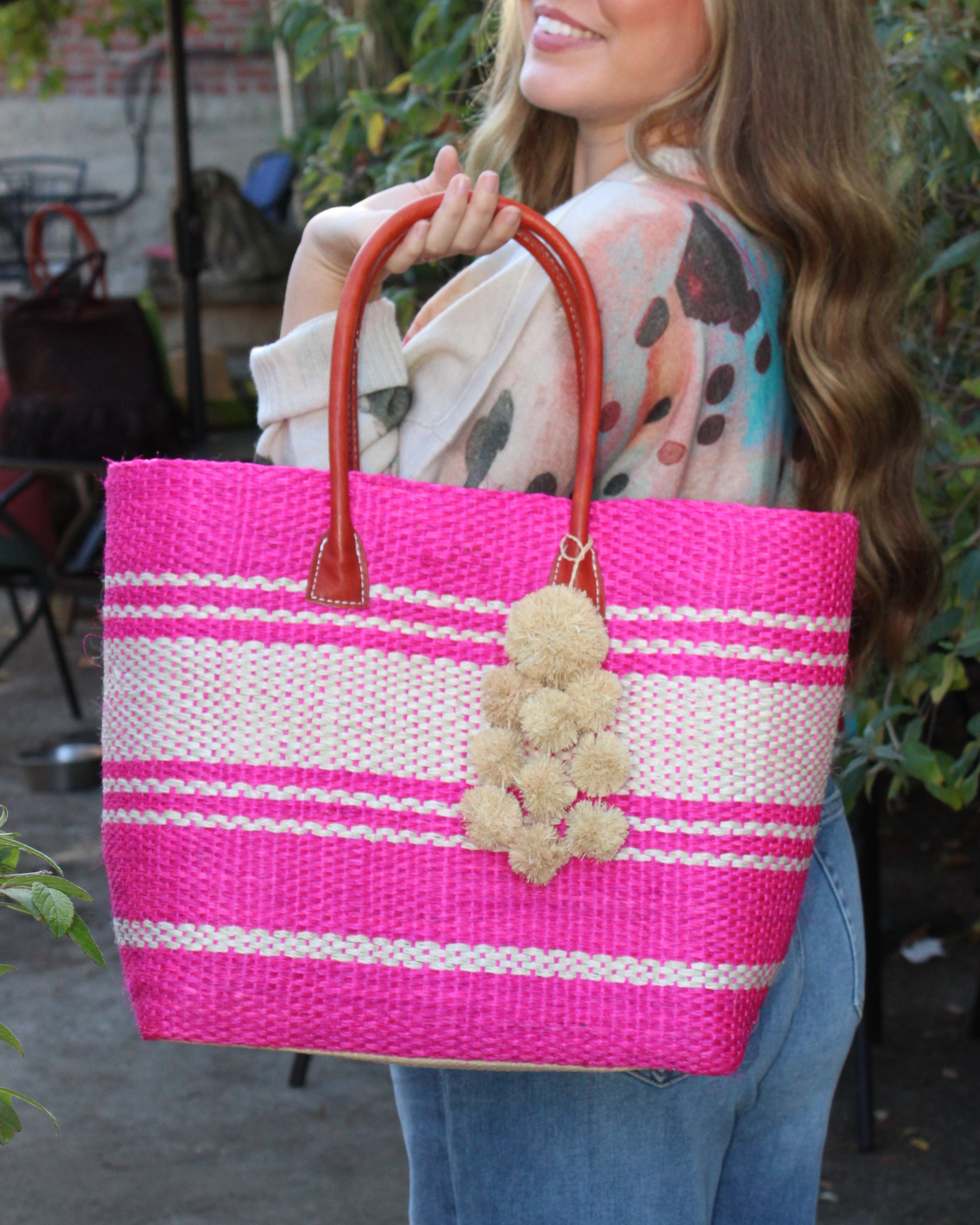 Model wearing Cabrillo Sisal Basket Bag with Waterfall Pompoms Charm Embellishment handmade natural sisal fiber woven into a multicolor two tone Fuchsia hot/bright/barbie/pink and natural straw color horizontal stripe pattern with muti-width bands of color plus leather handles and natural raffia charm shoulder bag beach bag - Shebobo