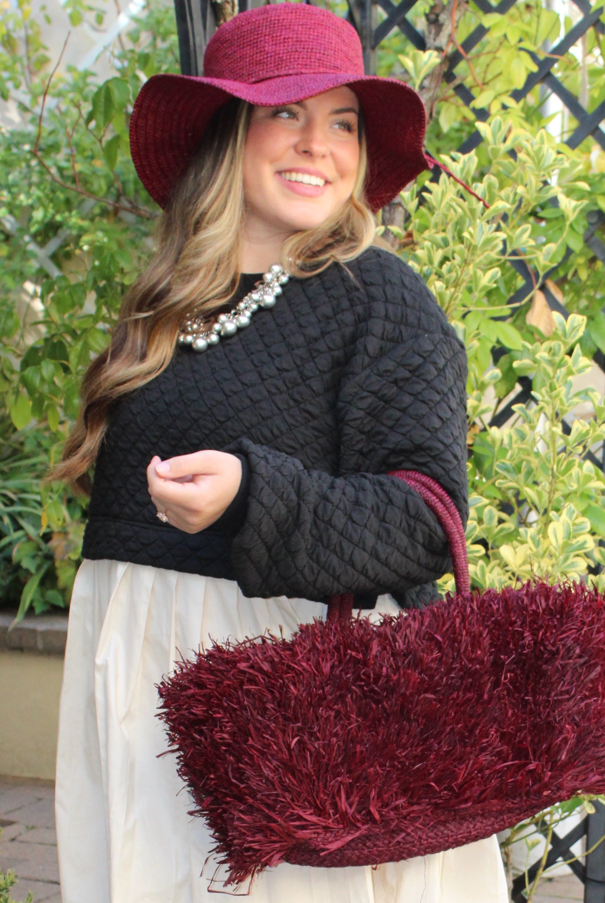 Model wearing Calypso Pompom Pouf Crochet Straw Basket Handbag handmade natural raffia palm fiber in a solid hue of Umberwine burgunday/bordeaux/dark/purple/red with brushed fringe strands crochet and knotted into a pouf texture with matching wrapped handles purse - Shebobo (with Bolero 3" Brim Crochet Straw Sun Hats)