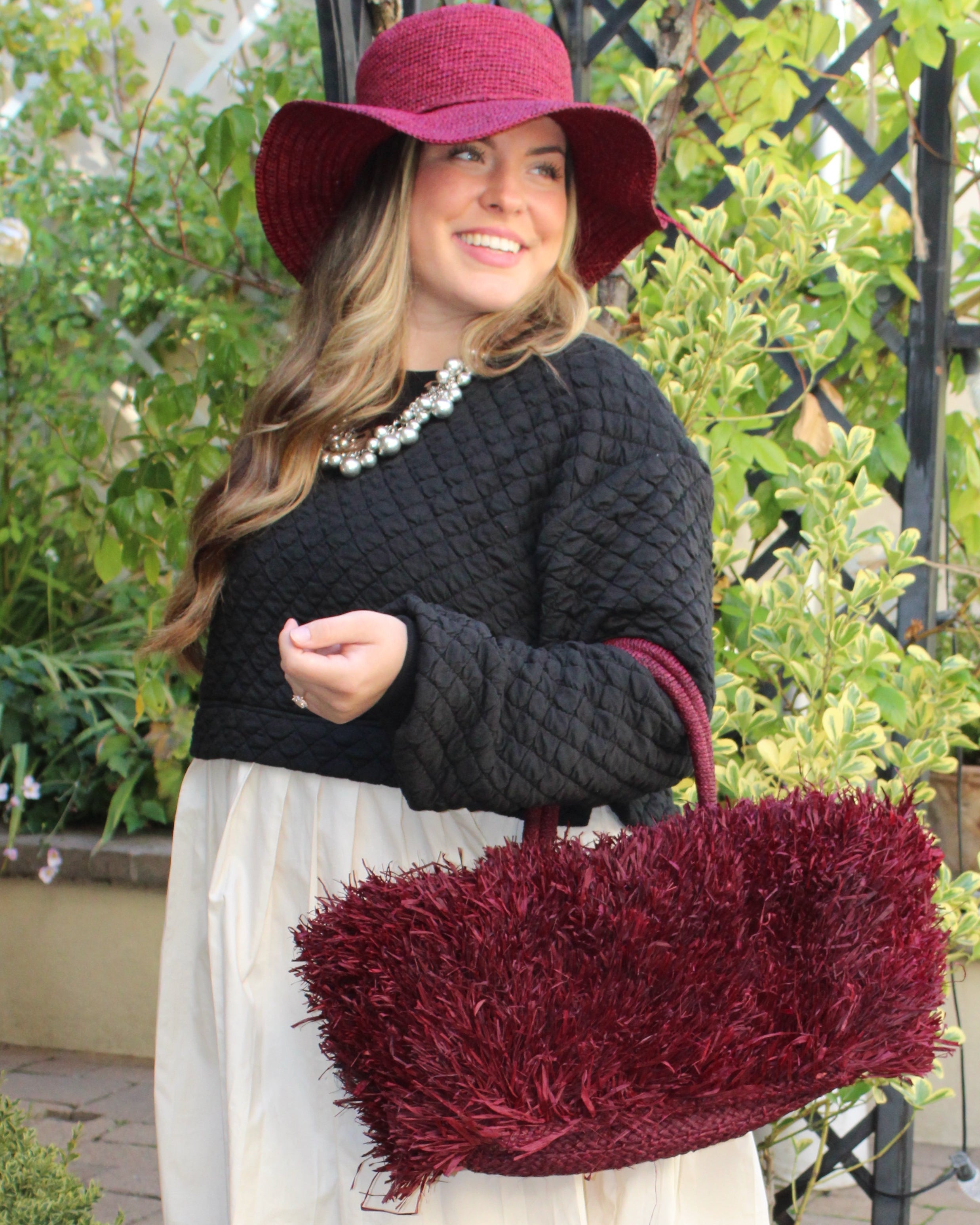 Model wearing Calypso Pompom Pouf Crochet Straw Basket Handbag handmade natural raffia palm fiber in a solid hue of Umberwine burgunday/bordeaux/dark/purple/red with brushed fringe strands crochet and knotted into a pouf texture with matching wrapped handles purse - Shebobo (with Bolero 3" Brim Crochet Straw Sun Hats)