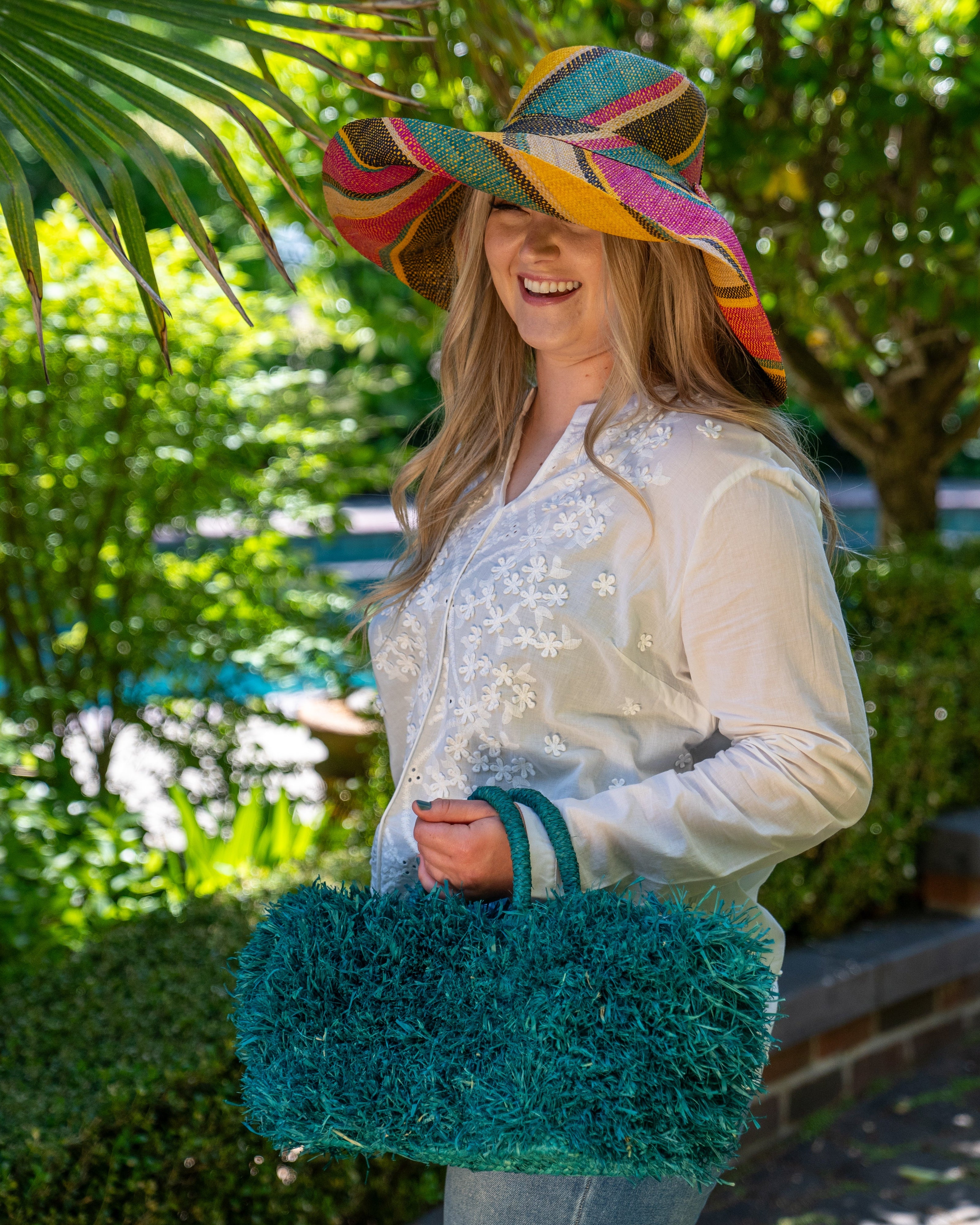 Model wearing Calypso Pompom Pouf Crochet Straw Basket Handbag handmade natural raffia palm fiber in a solid hue of Turquoise blue/green with brushed fringe strands crochet and knotted into a pouf texture with matching wrapped handles purse - Shebobo (with Monica 5" & 7" Wide Brim Multicolor Stripe Pattern Packable Straw Sun Hats)