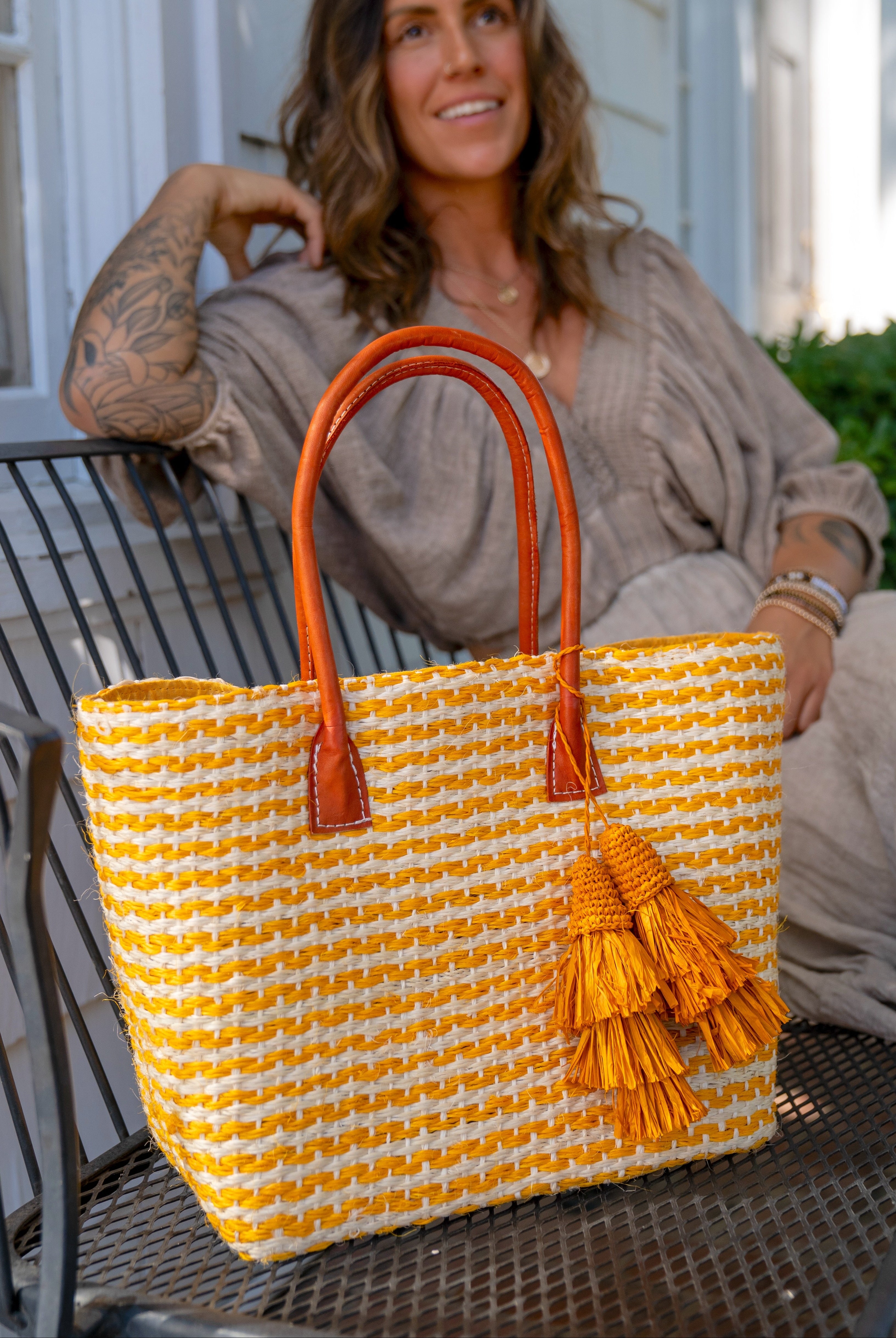 Model wearing Cancun Small Sisal Basket Bag with Tassel Charm handmade natural sisal fibers woven into even width horizontal bands of saffron yellow and natural straw color that create a pinstripe pattern with matching knotted raffia tufts tassel charm embellishment and leather handles beach bag - Shebob