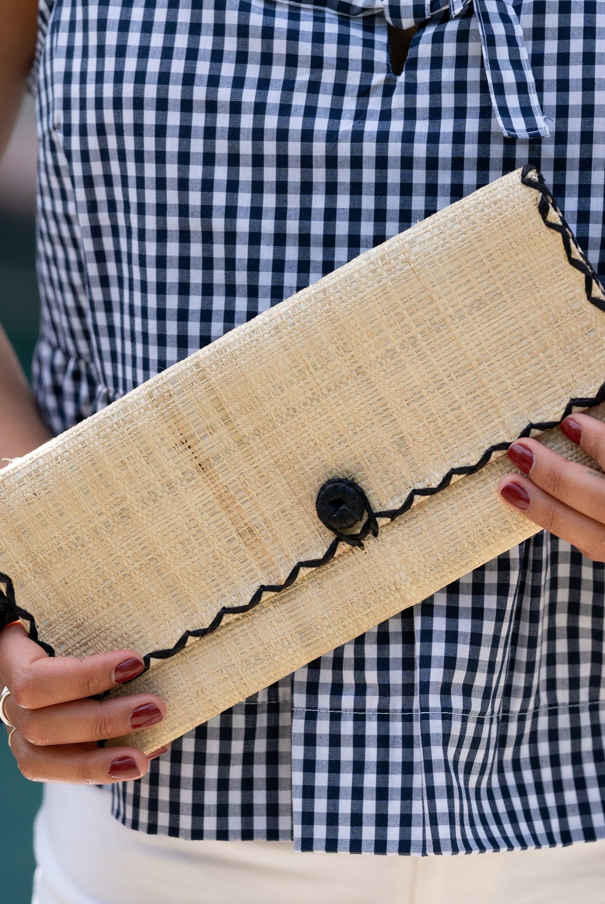 Model wearing ChiChi Natural Straw Clutch Bag handmade loomed raffia palm fiber wristlet in natural straw color with black cross stitch edging, button & loop closure, and braided wrist strap - Shebobo