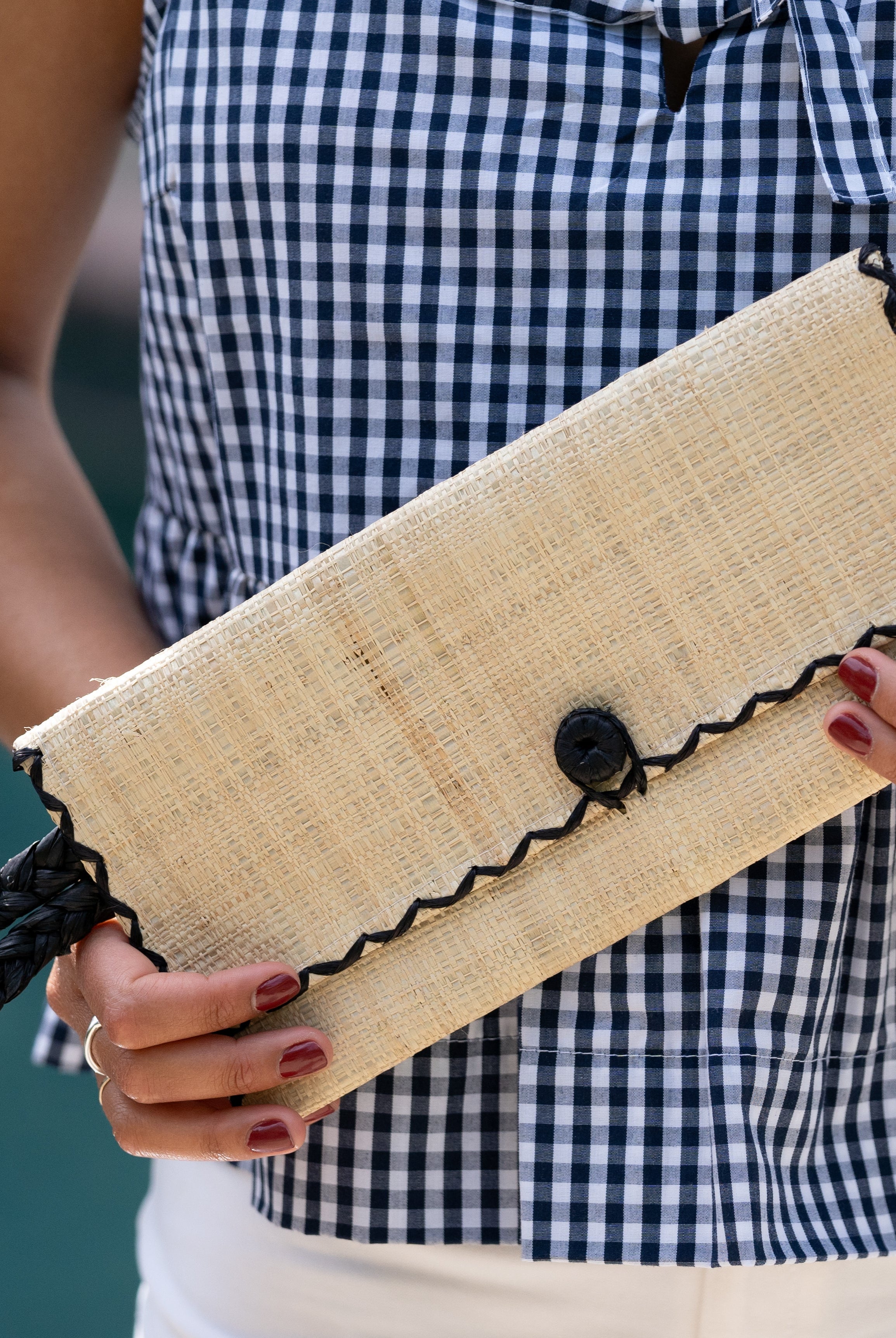 Model wearing ChiChi Natural Straw Clutch Bag handmade loomed raffia palm fiber wristlet in natural straw color with black cross stitch edging, button & loop closure, and braided wrist strap - Shebobo