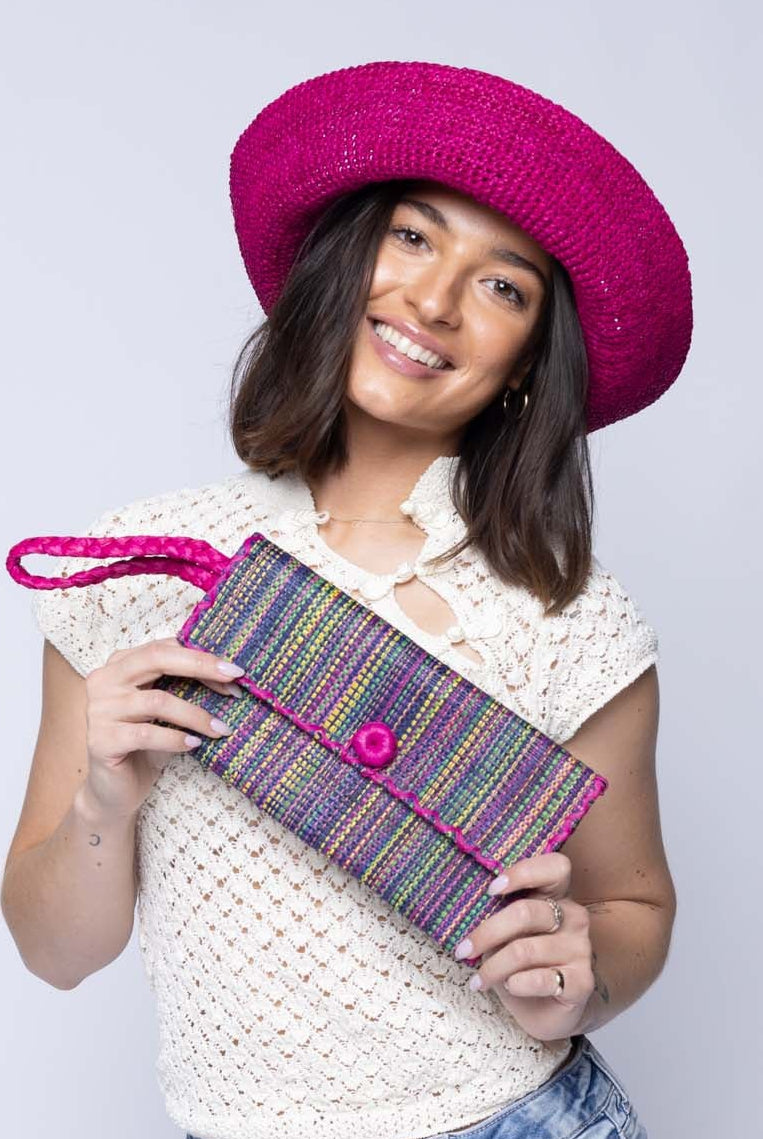 Model wearing ChiChi Multicolor Melange Multicolor Straw Clutch Bag handmade loomed raffia palm fiber wristlet in vertical heathered/blended stripe pattern of orchid pink/purple, navy dark/blue, fuchsia bright/pink, yellow, midori lime/green, light pink, etc. with matching color cross stitch edging, button & loop closure, and braided wrist strap - Shebobo
