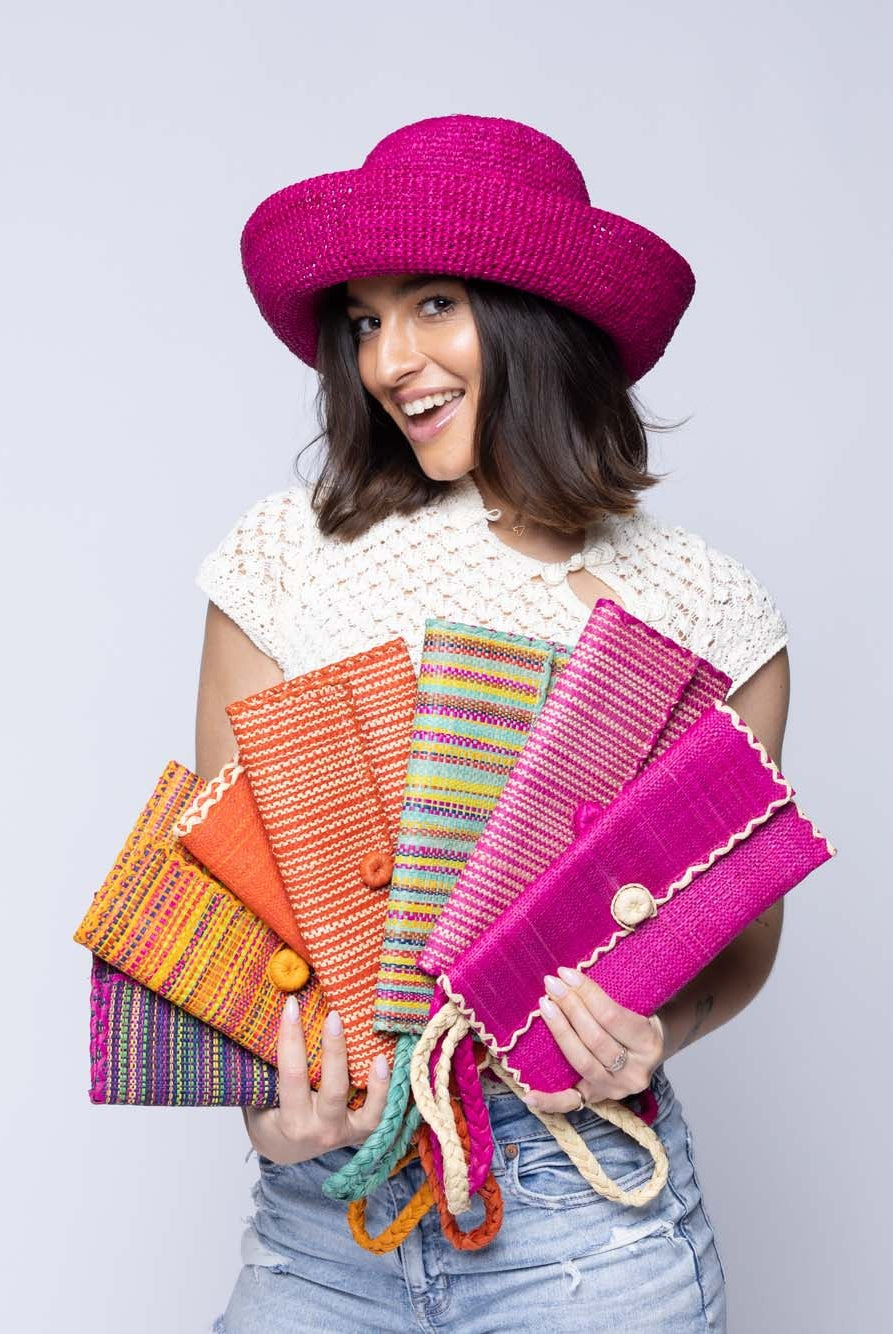 Model wearing multiple solid hues, two tone stripes, multicolor stripes, and melange patterns of ChiChi Straw Clutch Wristlet Bag handmade loomed raffia palm fiber in vibrant colors with natural cross stitch edging and raffia loop button closure with matching braided wrist loop - Shebobo