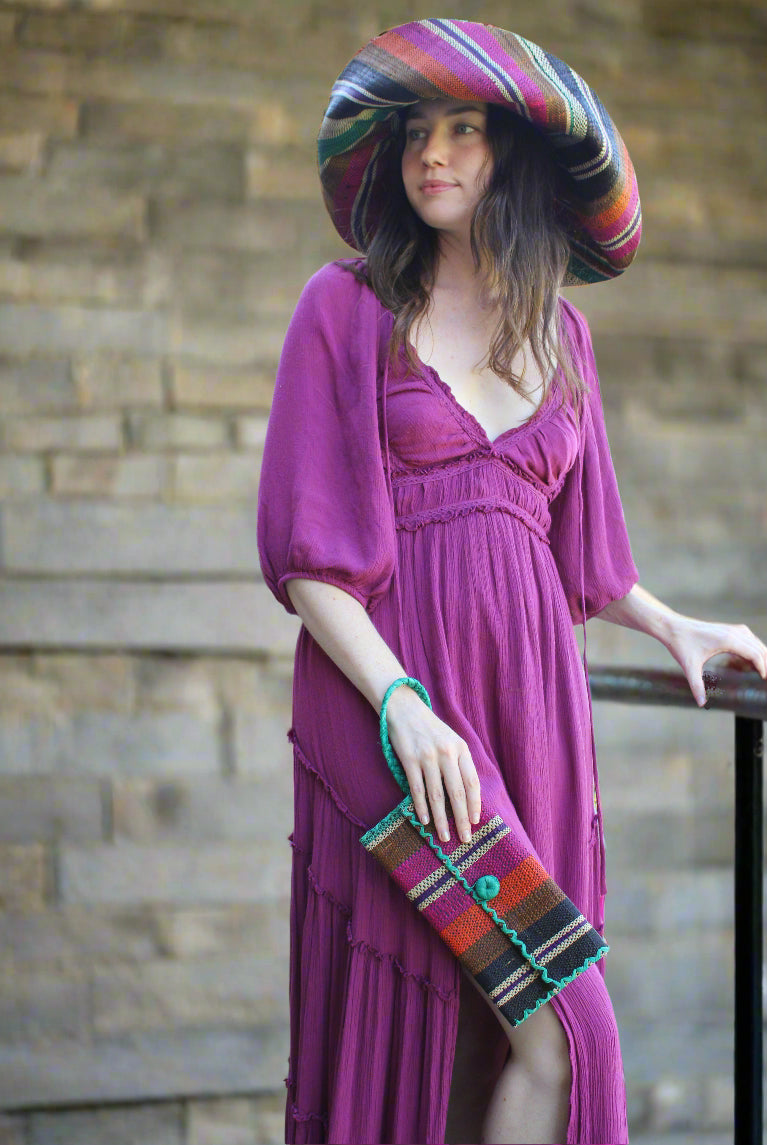 Model wearing ChiChi Revel Swirl Multicolor Straw Clutch Bag handmade loomed raffia palm fiber wristlet in multi-width bands of teal dark/blue/green, fuchsia hot/bright/barbie/pink, deep purple, black, coral orange/red, cinnamon tobacco/dark/brown, and natural straw color that create a vertical stripe pattern with teal color cross stitch edging, button & loop closure, and braided wrist strap - Shebobo (with Monica 5" & 7" Wide Brim Multicolor Stripe Pattern Packable Straw Sun Hats)