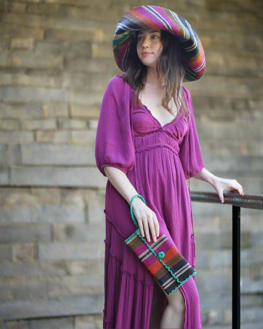 Model wearing ChiChi Revel Swirl Multicolor Straw Clutch Bag handmade loomed raffia palm fiber wristlet in multi-width bands of teal dark/blue/green, fuchsia hot/bright/barbie/pink, deep purple, black, coral orange/red, cinnamon tobacco/dark/brown, and natural straw color that create a vertical stripe pattern with teal color cross stitch edging, button & loop closure, and braided wrist strap - Shebobo (with Monica 5" & 7" Wide Brim Multicolor Stripe Pattern Packable Straw Sun Hats)