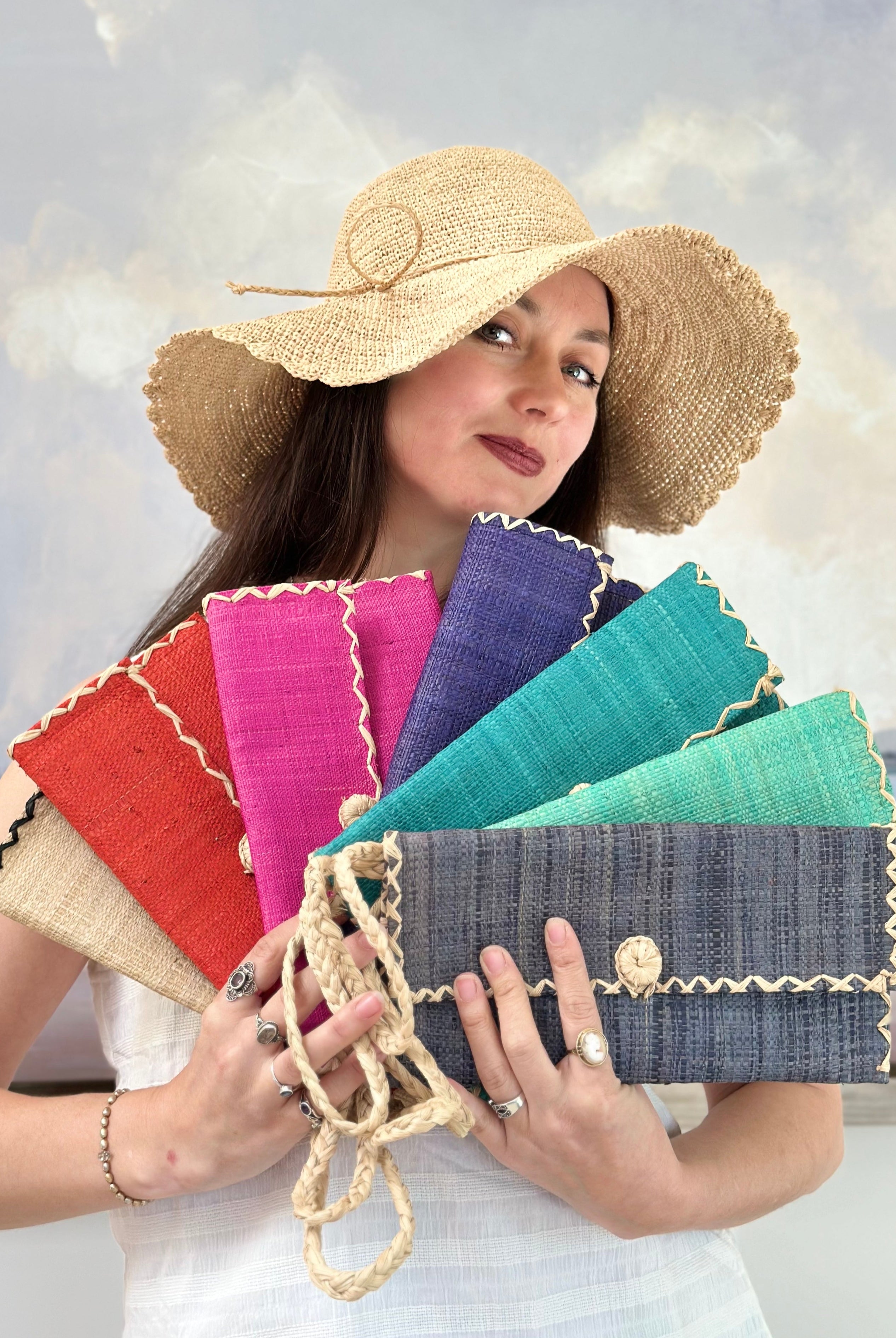 Model wearing multiple solid hues of ChiChi Straw Clutch Wristlet Bag handmade loomed raffia palm fiber in vibrant colors with natural cross stitch edging and raffia loop button closure with matching braided wrist loop with 4" Brim Genevieve Crochet Straw Sun Hats with Lace Weave Edge in Natural Raffia - Shebobo
