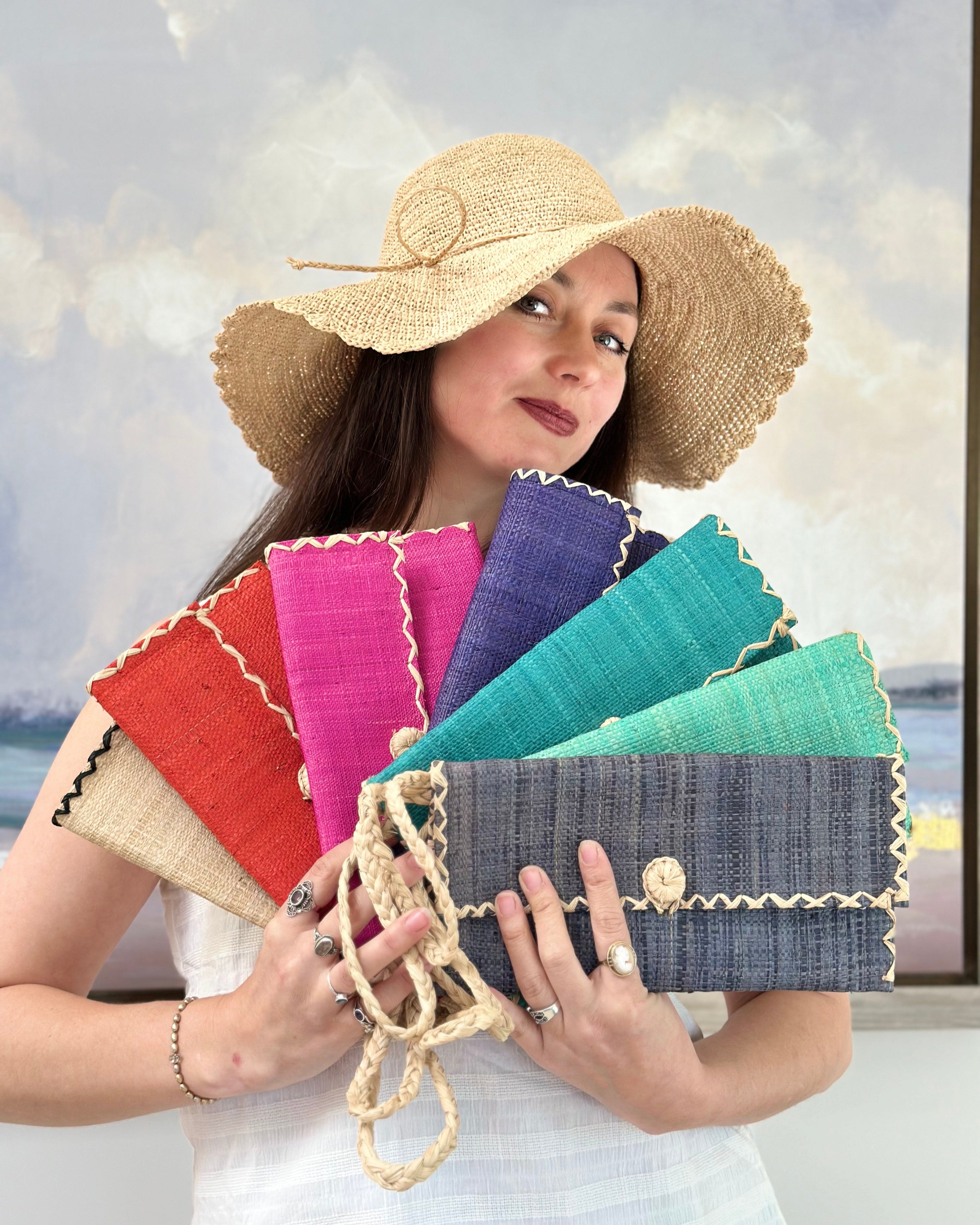 Model wearing multiple solid hues of ChiChi Straw Clutch Wristlet Bag handmade loomed raffia palm fiber in vibrant colors with natural cross stitch edging and raffia loop button closure with matching braided wrist loop with 4" Brim Genevieve Crochet Straw Sun Hats with Lace Weave Edge in Natural Raffia - Shebobo