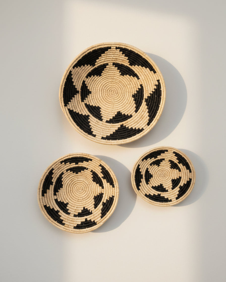 Life style view Dakarai Raffia Straw Wall Decor Display Basket Tray: Set of 3 handmade woven natural raffia palm fiber in a crochet loop technique concentric coiled circular pattern with double natural straw color and black centered star flower pattern set of three sized flat bowl trays - Shebobo