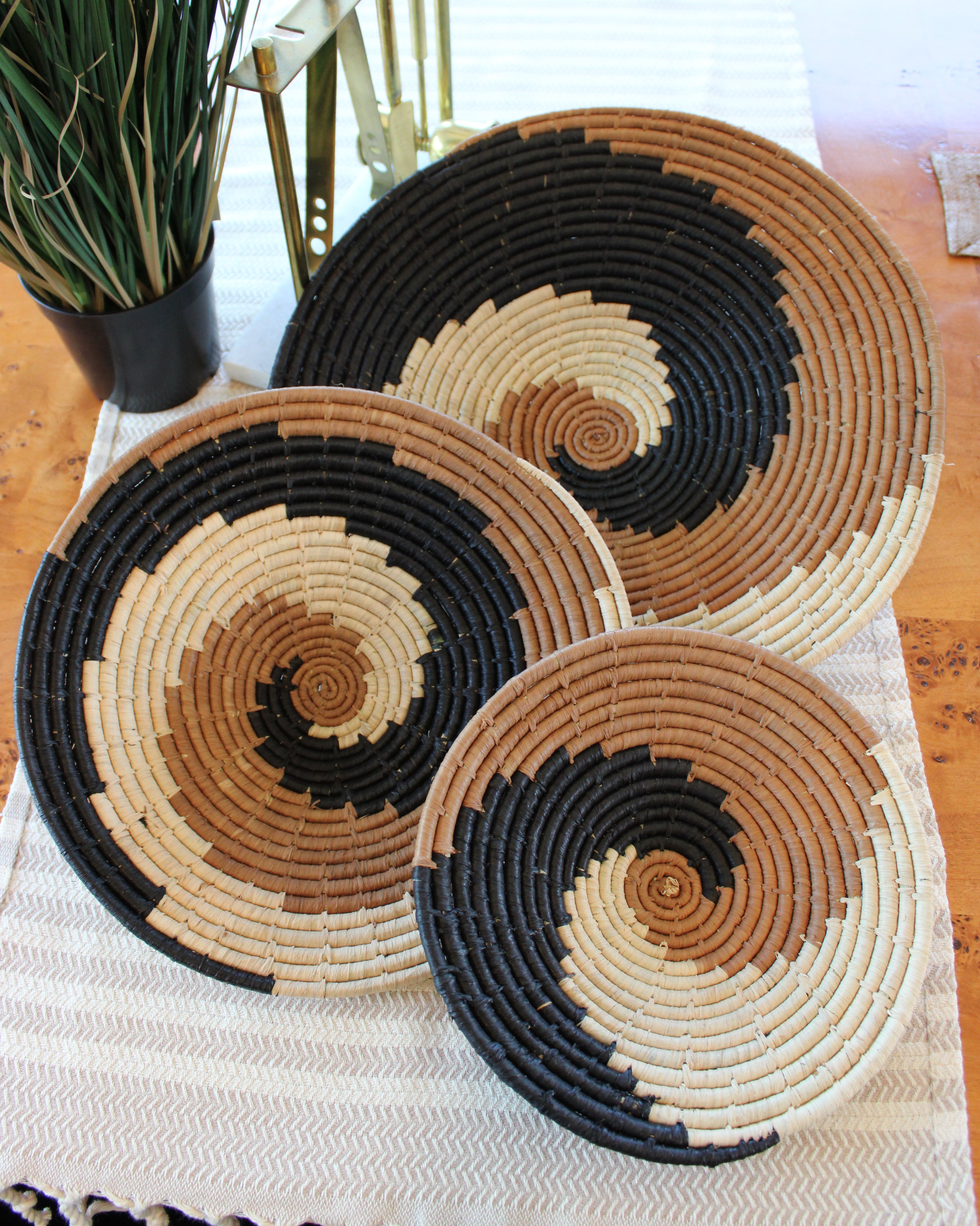 Home decor lifestyle view Dakarai Raffia Straw Wall Decor Display Basket Trays: Set of 3 handmade woven raffia palm fiber in a crochet loop technique creating a concentric swirl pattern of bands of natural straw color, cinnamon/tobacco/brown, and black in a three tone spiral from the center to the outer edge set of three sized flat bowl trays - Shebobo