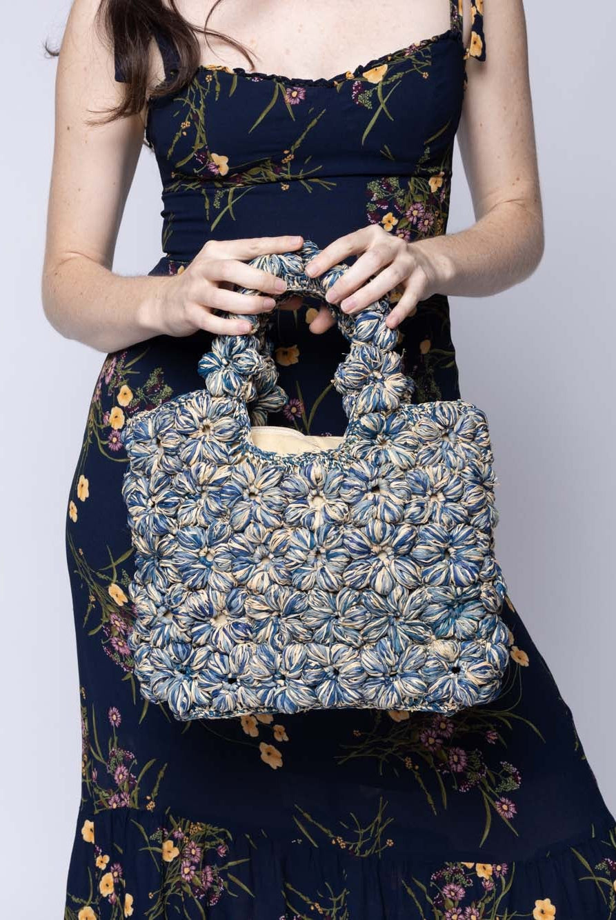 Model wearing Florence Denim Blues Multicolor Flower Crochet Straw Basket Handbag handmade natural raffia palm fiber in a multicolor hue combination of natural straw color, denim light/retro/blue, and luminous navy/dark/blue with subtle tied flower design consisting of 88 flowers crochet together along the outside of the bag as well as the handles purse tote - Shebobo