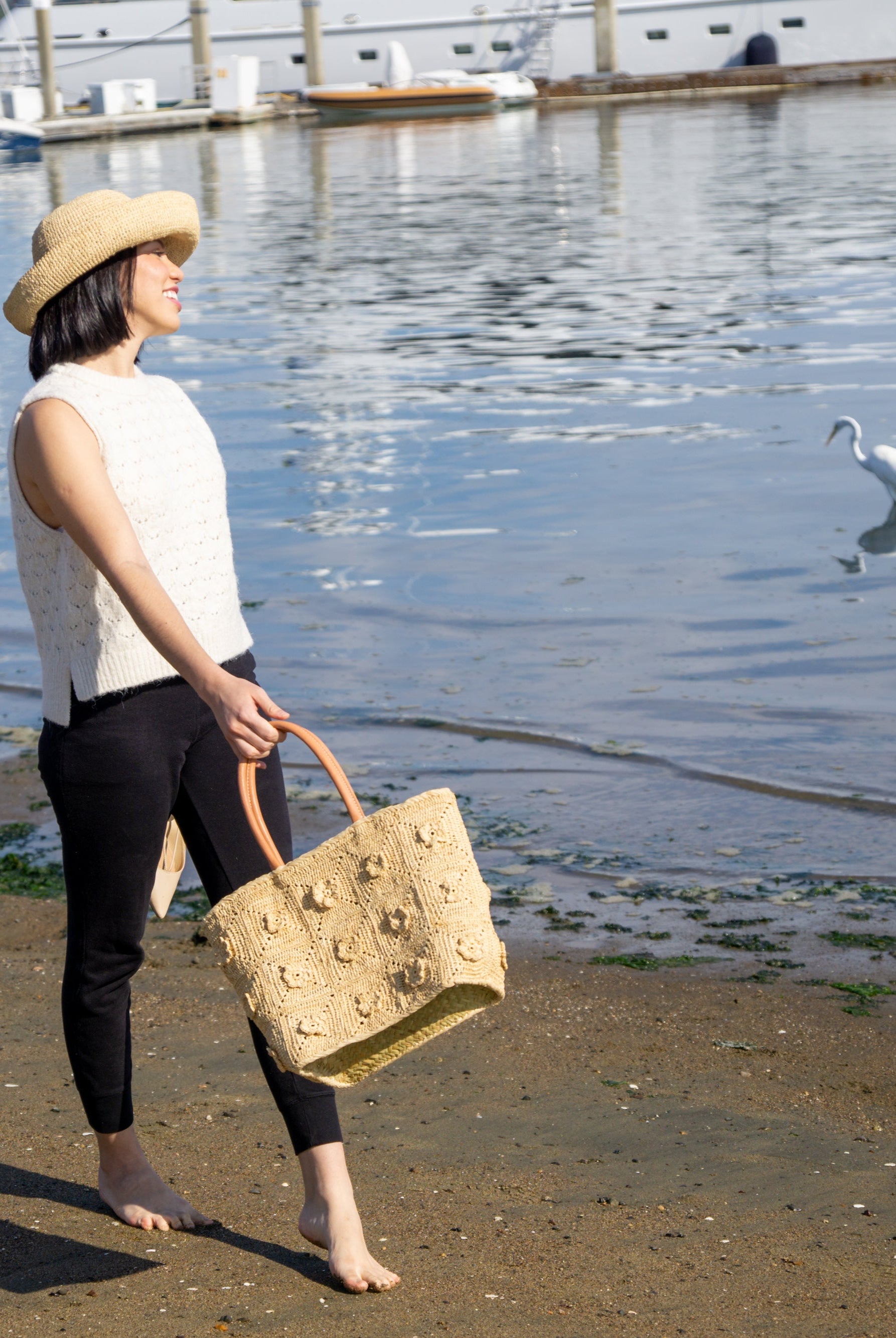 Model wearing Flower Power Natural Crochet Straw Basket Bag handmade from natural raffia palm fiber in a granny square-esque geometric floral pattern with flower detailing projecting from main crochet textile in a natural straw color with leather handles handbag purse - Shebobo (with Leor 3" Natural Crochet Straw Sun Hat)