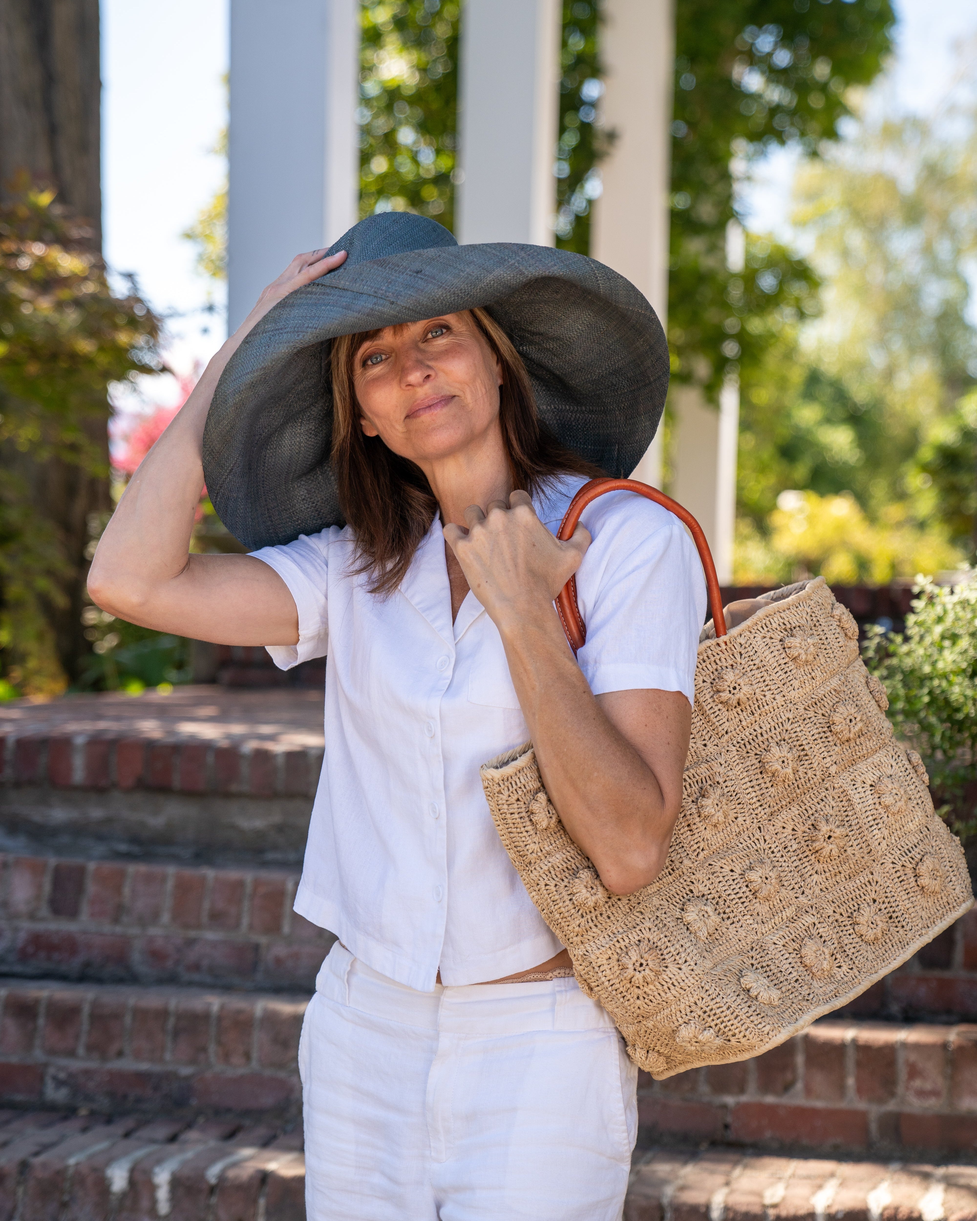 Model wearing Flower Power Natural Crochet Straw Basket Bag handmade from natural raffia palm fiber in a granny square-esque geometric floral pattern with flower detailing projecting from main crochet textile in a natural straw color with leather handles handbag purse - Shebobo (with Monica 5" & 7" Wide Brim Solid Color Packable Straw Sun Hats)