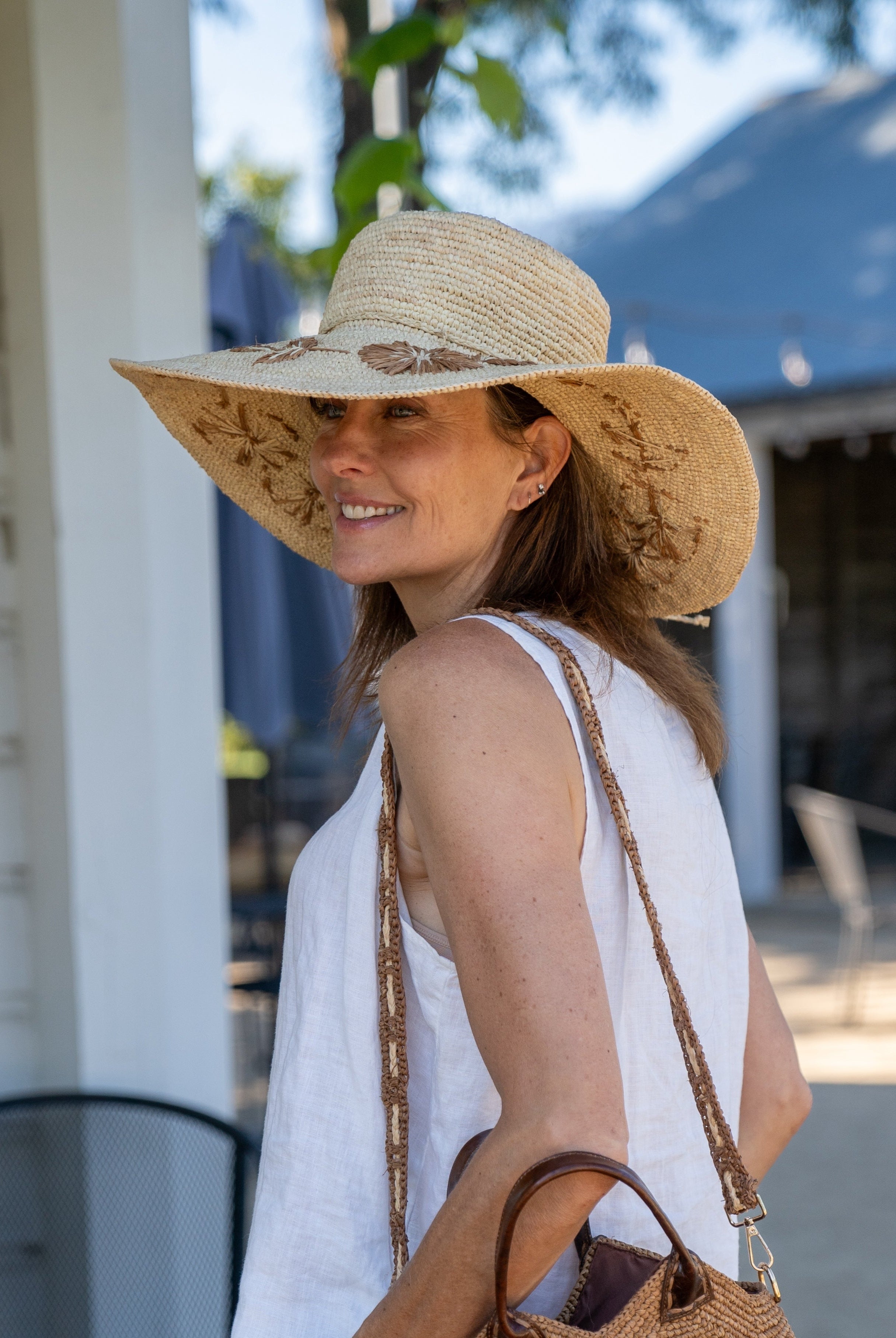 Model wearing 5.5" Brim Gigi Natural and Cinnamon Floral Embroidered Crochet Straw Sun Hats handmade crochet natural raffia palm fiber in a solid hue of natural straw color making a concentric pattern from the center top of the flattened crown spiraling out to the edge of the brim with matching adjustable raffia braid hat band and cinnamon dark brown floral pattern with leaves embroidered on the brim - Shebobo