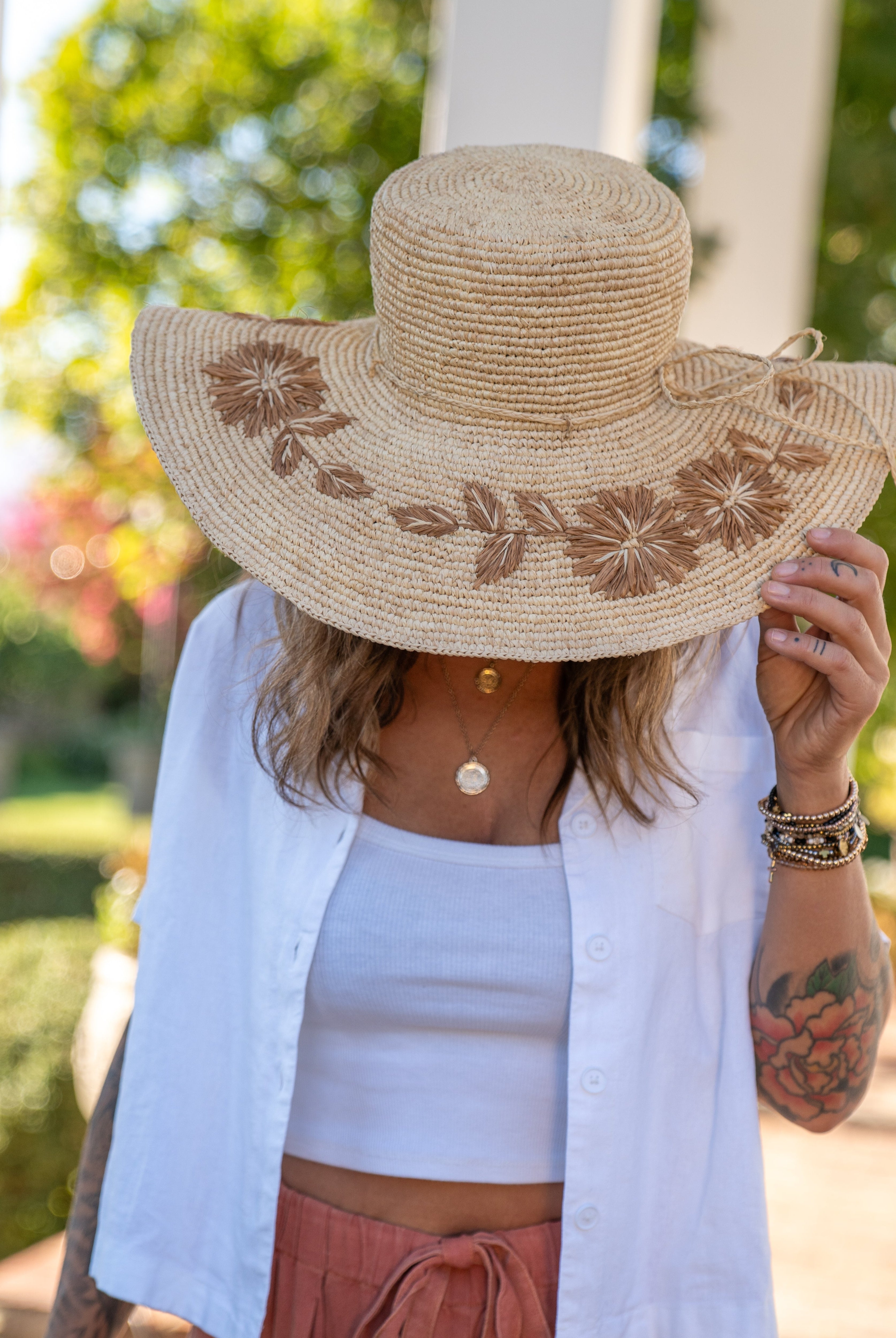 Model wearing 5.5" Brim Gigi Natural and Cinnamon Floral Embroidered Crochet Straw Sun Hats handmade crochet natural raffia palm fiber in a solid hue of natural straw color making a concentric pattern from the center top of the flattened crown spiraling out to the edge of the brim with matching adjustable raffia braid hat band and cinnamon dark brown floral pattern with leaves embroidered on the brim - Shebobo
