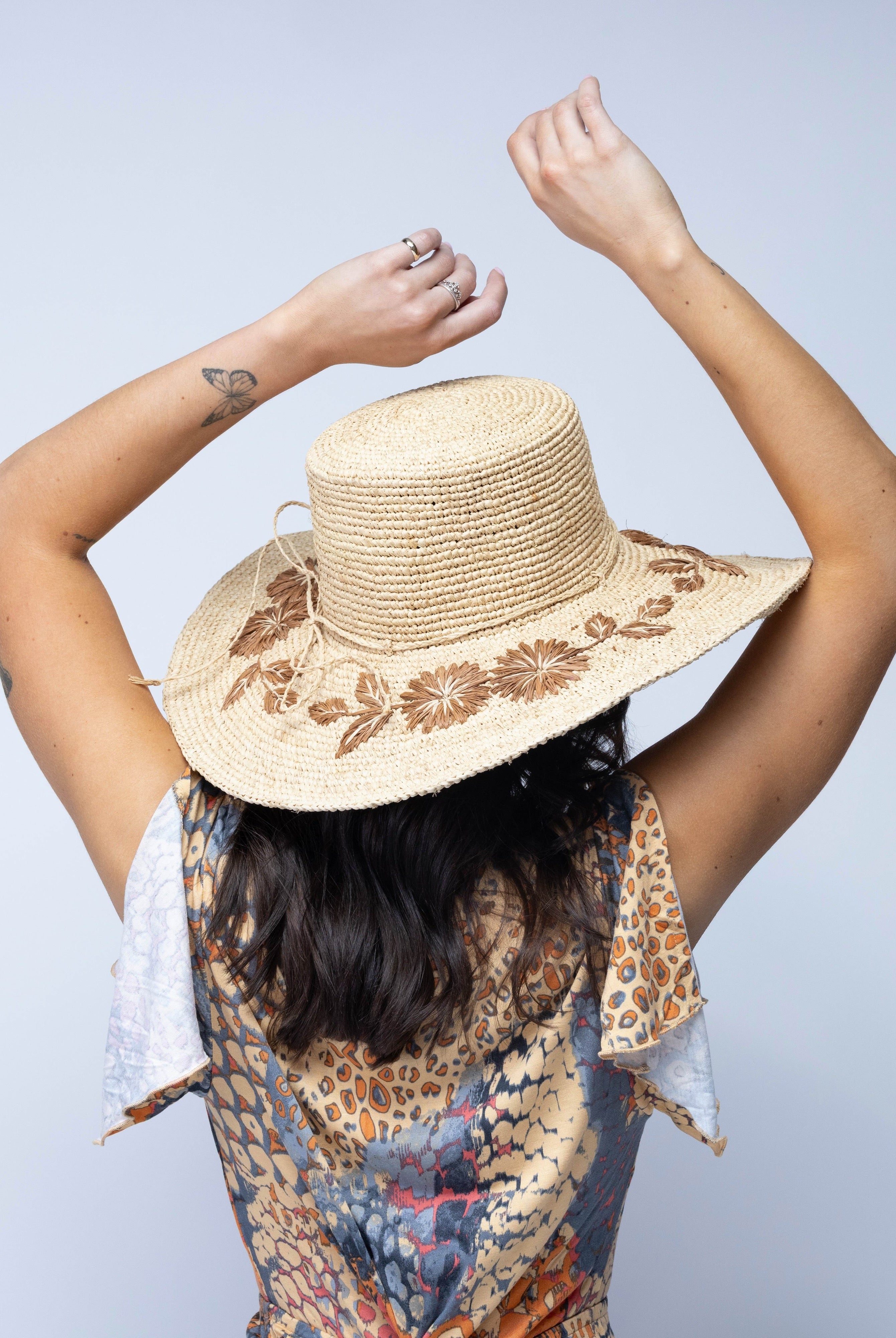 Model wearing 5.5" Brim Gigi Natural and Cinnamon Floral Embroidered Crochet Straw Sun Hats handmade crochet natural raffia palm fiber in a solid hue of natural straw color making a concentric pattern from the center top of the flattened crown spiraling out to the edge of the brim with matching adjustable raffia braid hat band and cinnamon dark brown floral pattern with leaves embroidered on the brim - Shebobo