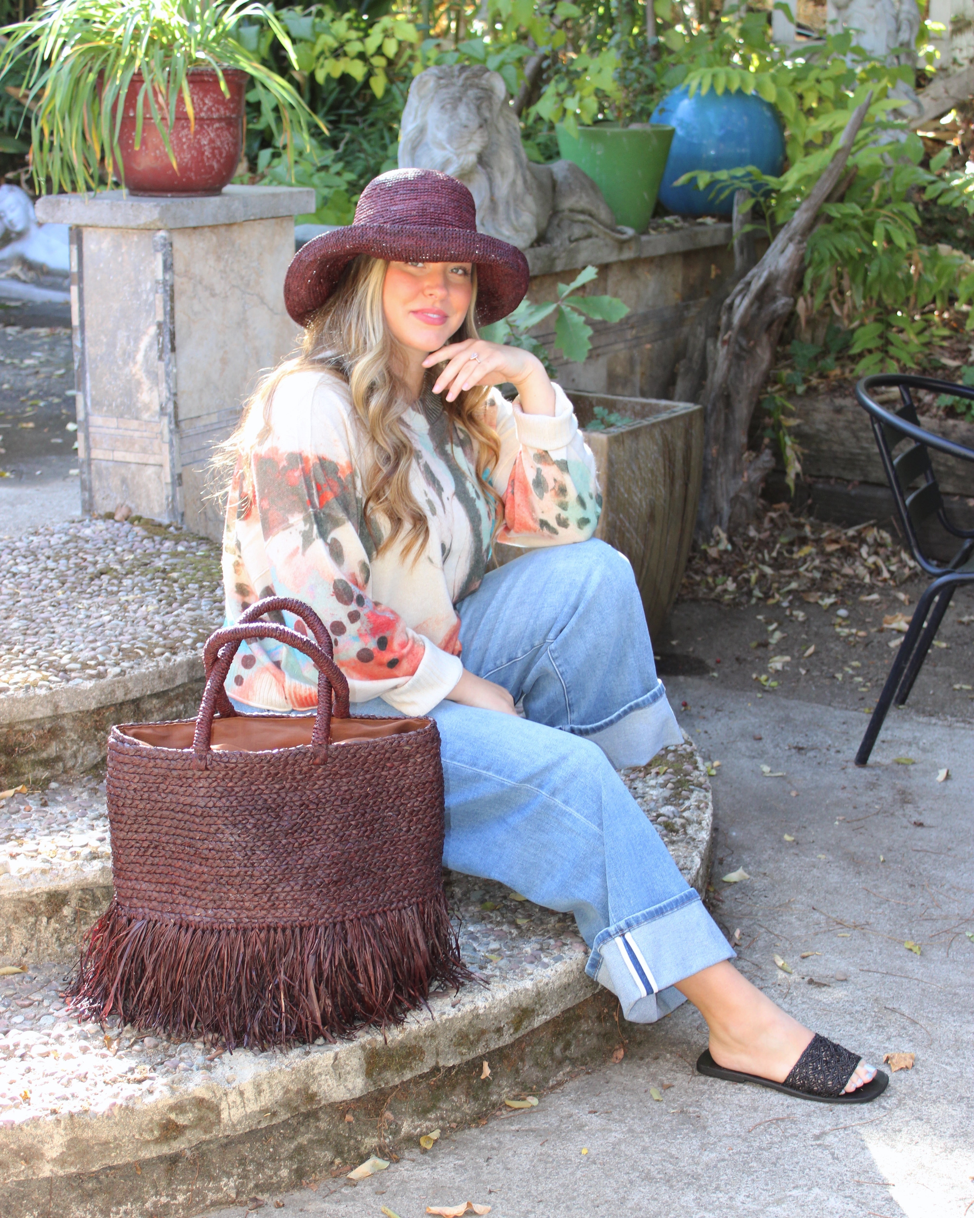 Model wearing Hanalei Braided Straw Basket Shoulder Bag with Fringe handmade braided natural raffia palm fiber in a solid hue of Mocha chocolate/dark/brown with horizontal bands of braids making a subtle texture and knotted long brushed fringe band around the botom third of the bag and matching looped handles beach bag purse - Shebobo (with Leor 3" Brim Crochet Straw Sun Hats & Lulu Macrame and Crochet Diamond Straw Sandal Shoes)