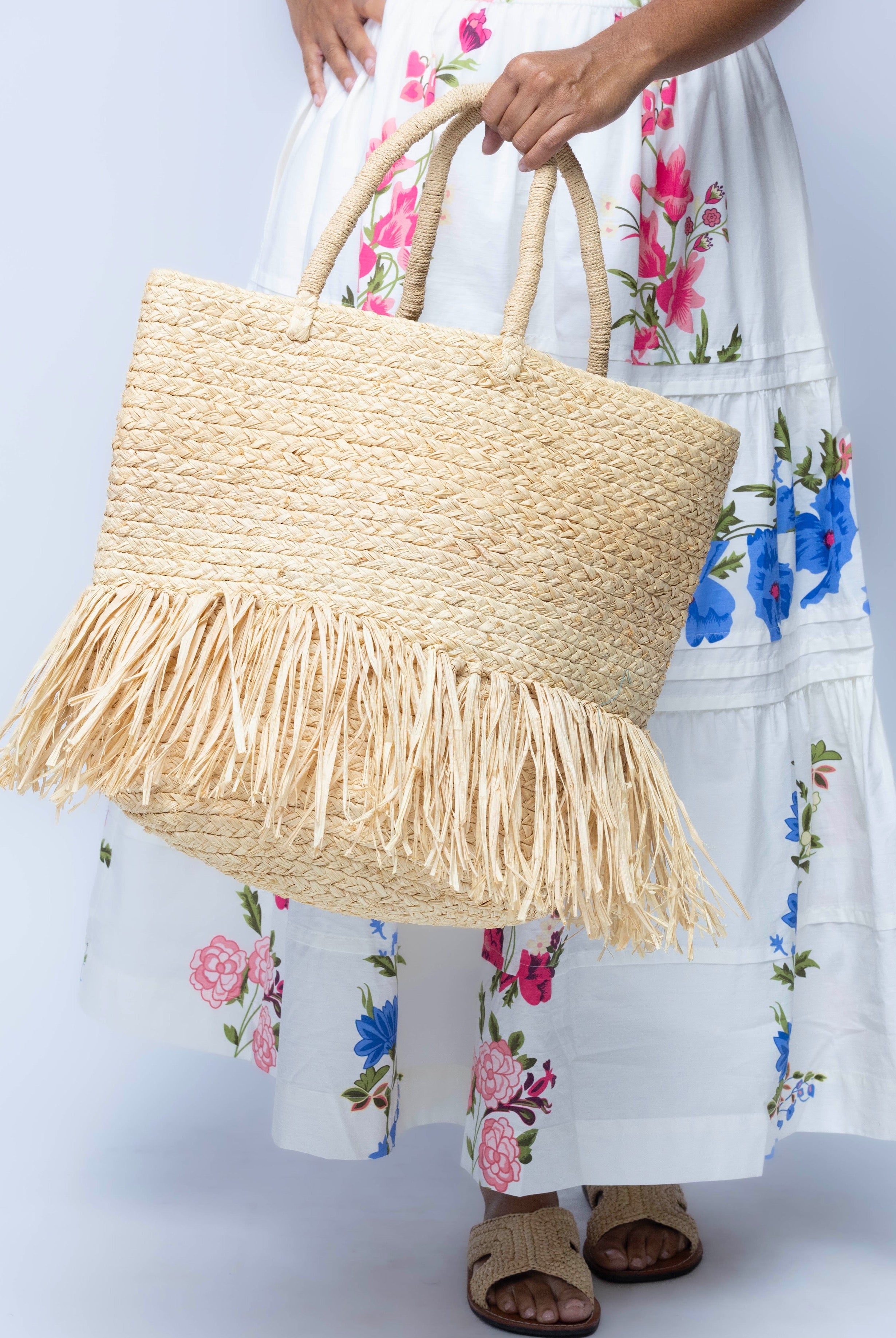 Model wearing Hanalei Braided Straw Basket Shoulder Bag with Fringe handmade braided natural raffia palm fiber in a solid hue of Natural straw color with horizontal bands of braids making a subtle texture and knotted long brushed fringe band around the botom third of the bag and matching looped handles beach bag purse - Shebobo