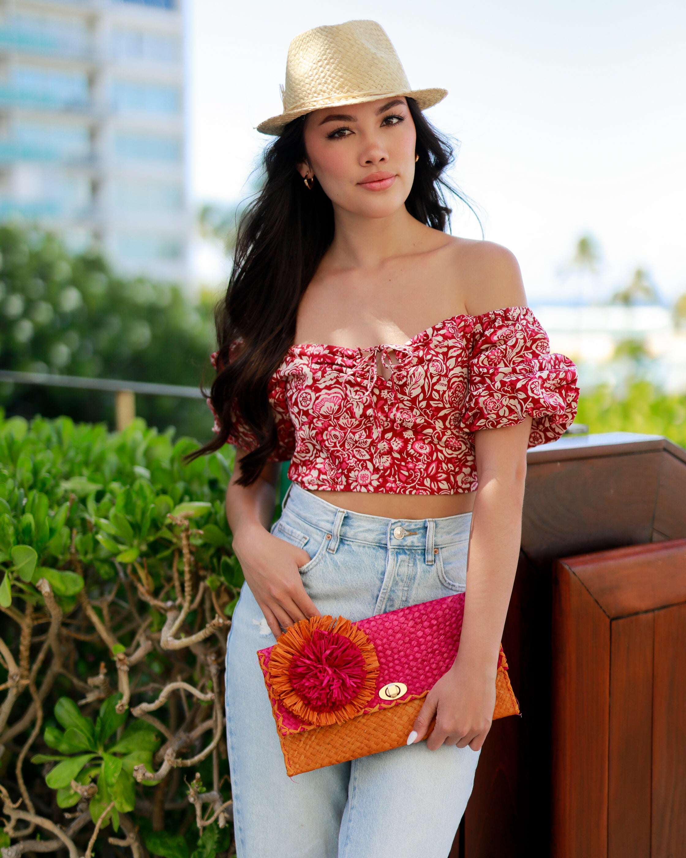 Model wearing Holden Ombre Fuchsia Straw Clutch with Flower Pouf handmade woven raffia dip dyed gradient of fuchsia bright/barbie/pink, saffron yellow, and coral orange/red with large coral and fuchsia fringe flower embellishment - Shebobo (with Charlie Natural Unisex Fedora Straw Hat)